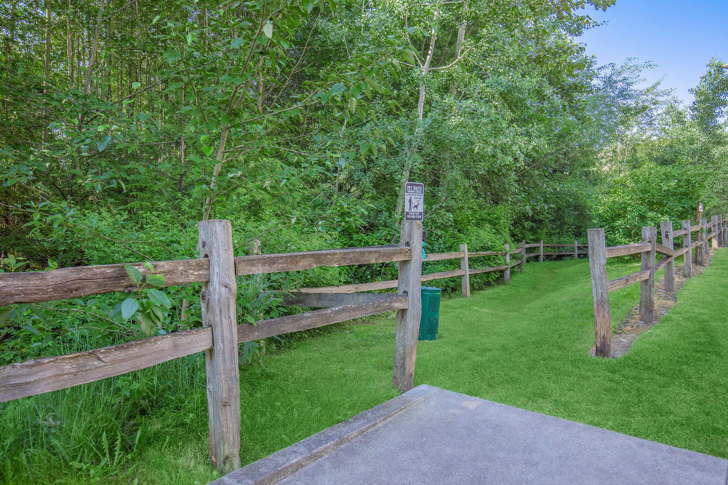 A serene pathway lined with wooden fencing, surrounded by lush greenery. The path leads into a vibrant natural area, with trees and bushes on either side. A sign is visible along the fence, indicating a designated area for pets. The scene conveys a tranquil outdoor environment for walking or recreational activities.