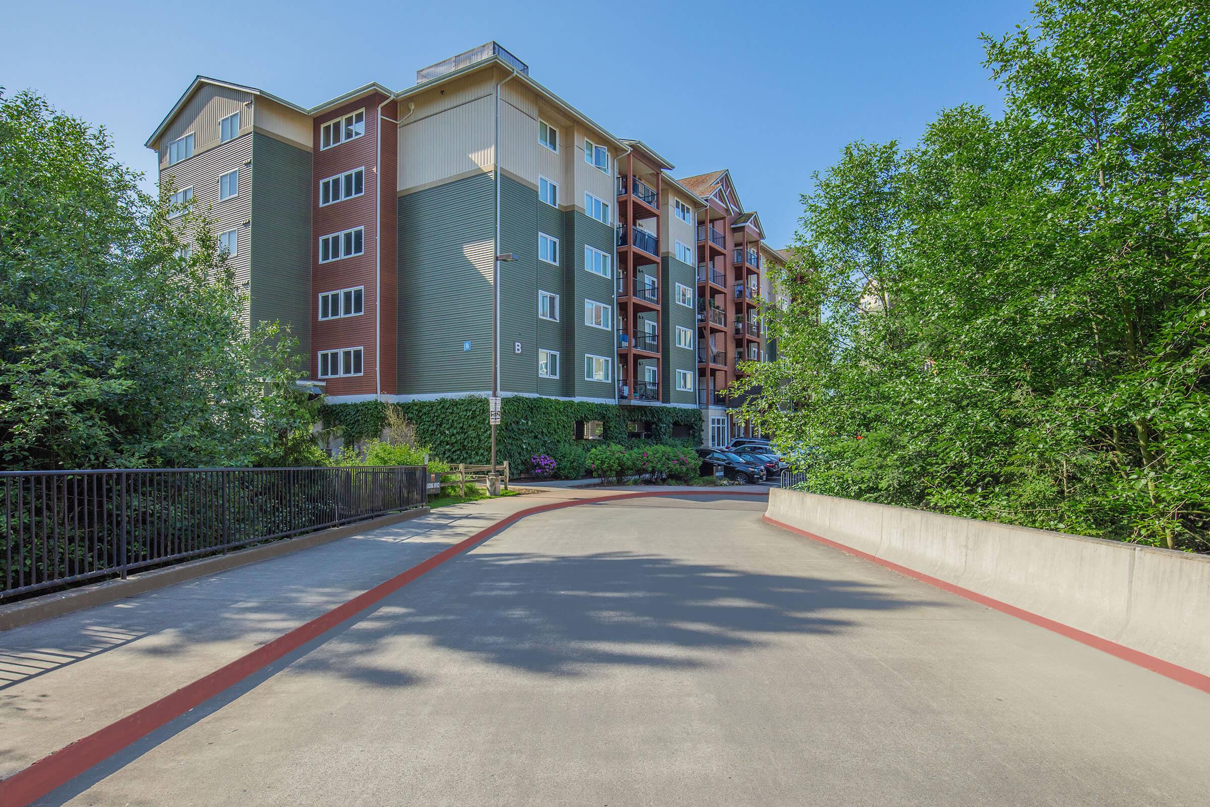 A view of a modern multi-story apartment building surrounded by greenery. The image shows a paved driveway leading to the entrance, with shrubs and trees lining the path. Clear blue skies are visible overhead, enhancing the inviting atmosphere of the residential area.
