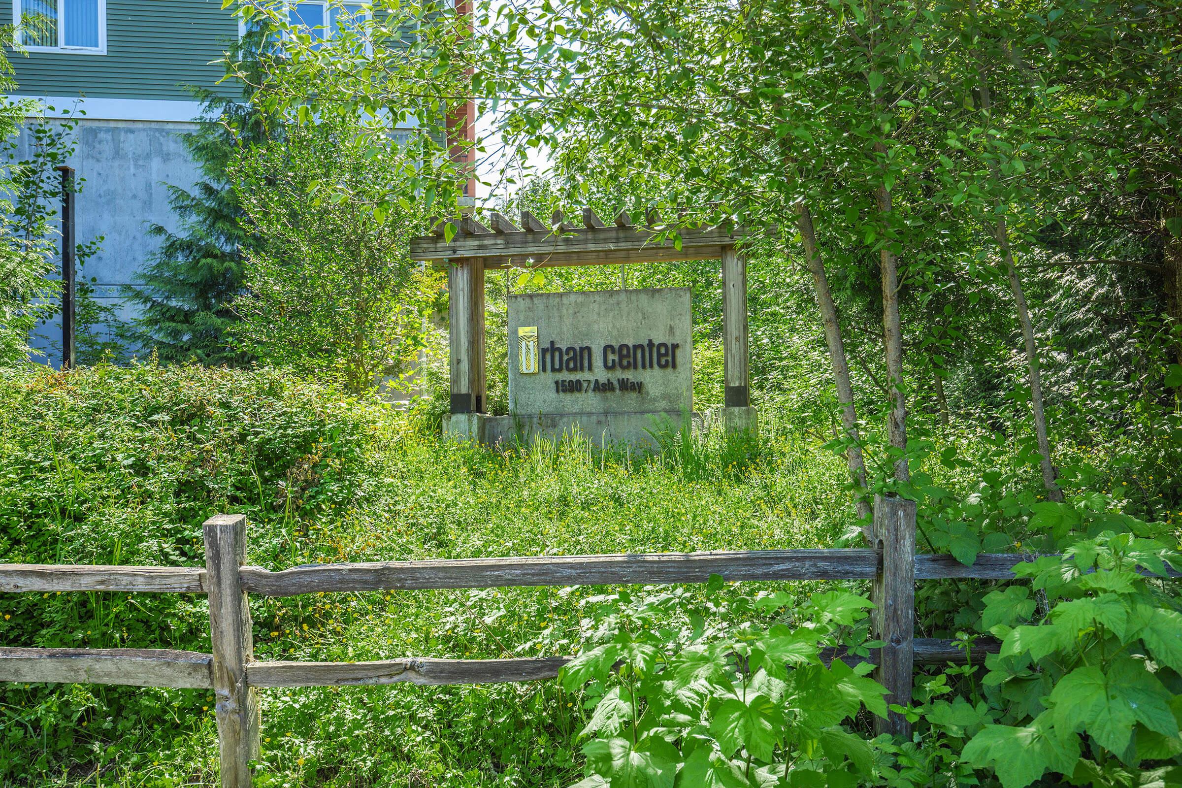 A sign for "Urban Center" surrounded by lush greenery and bushes, with a wooden fence in the foreground. The background features a hint of a building partially visible through the foliage.