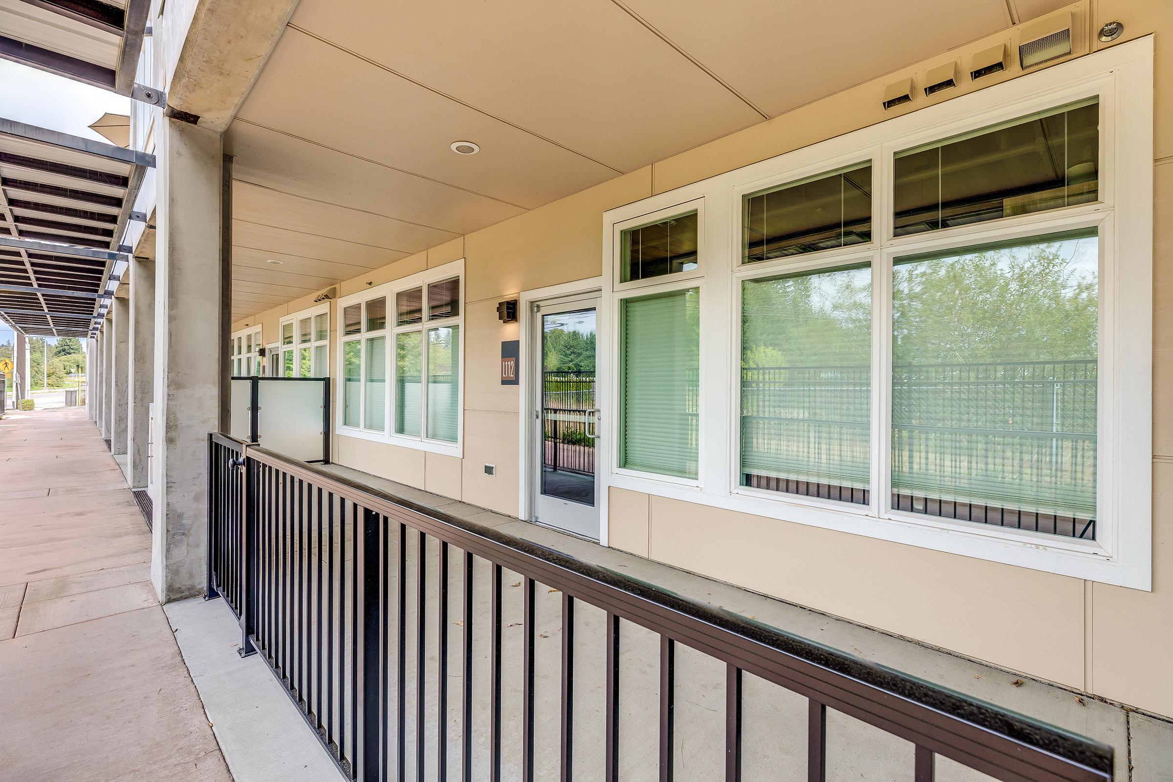 A corridor of a modern building featuring large windows, a gray concrete floor, and a black metal railing. The walls are light-colored, and there are greenery and outdoor spaces visible through the windows. The overall design appears contemporary and functional.