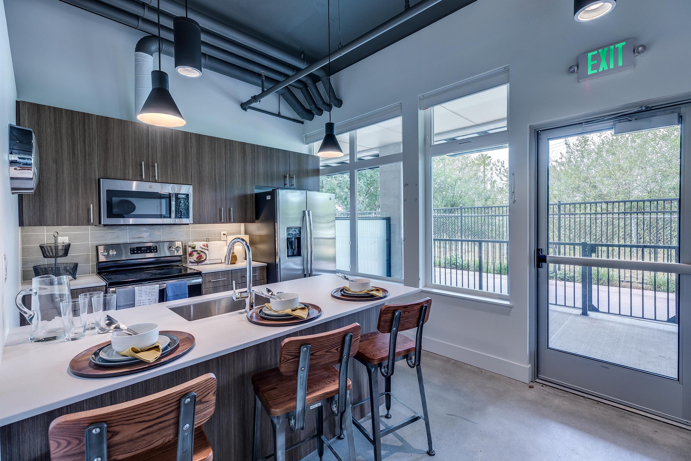 Modern kitchen featuring stainless steel appliances, a countertop with three wooden stools, and natural light coming from large windows. The decor includes dark wood cabinetry and contemporary lighting. An exit door leads to an outdoor area.