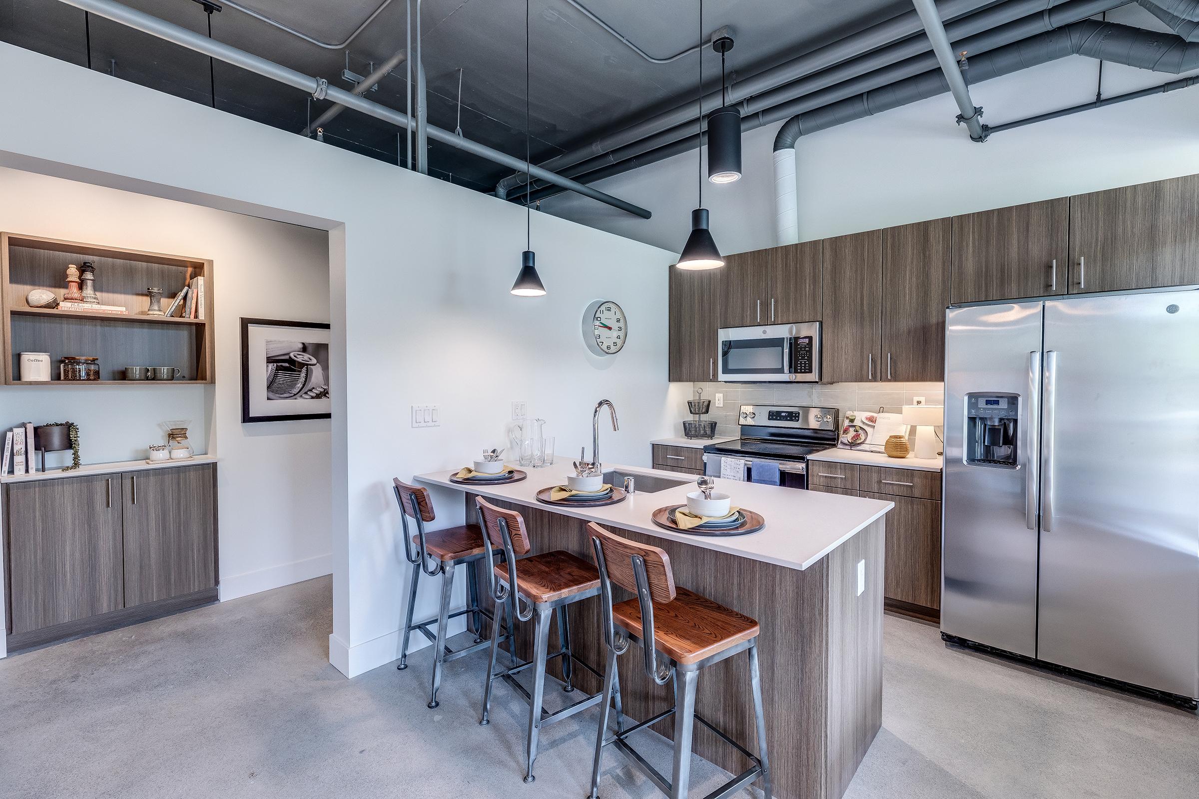 Modern kitchen featuring a central island with bar stools, stainless steel appliances, gray cabinetry, and pendant lighting. A clock is mounted on the wall, and there are decorative items on shelves in the background. The space has a clean, contemporary design with an industrial touch.