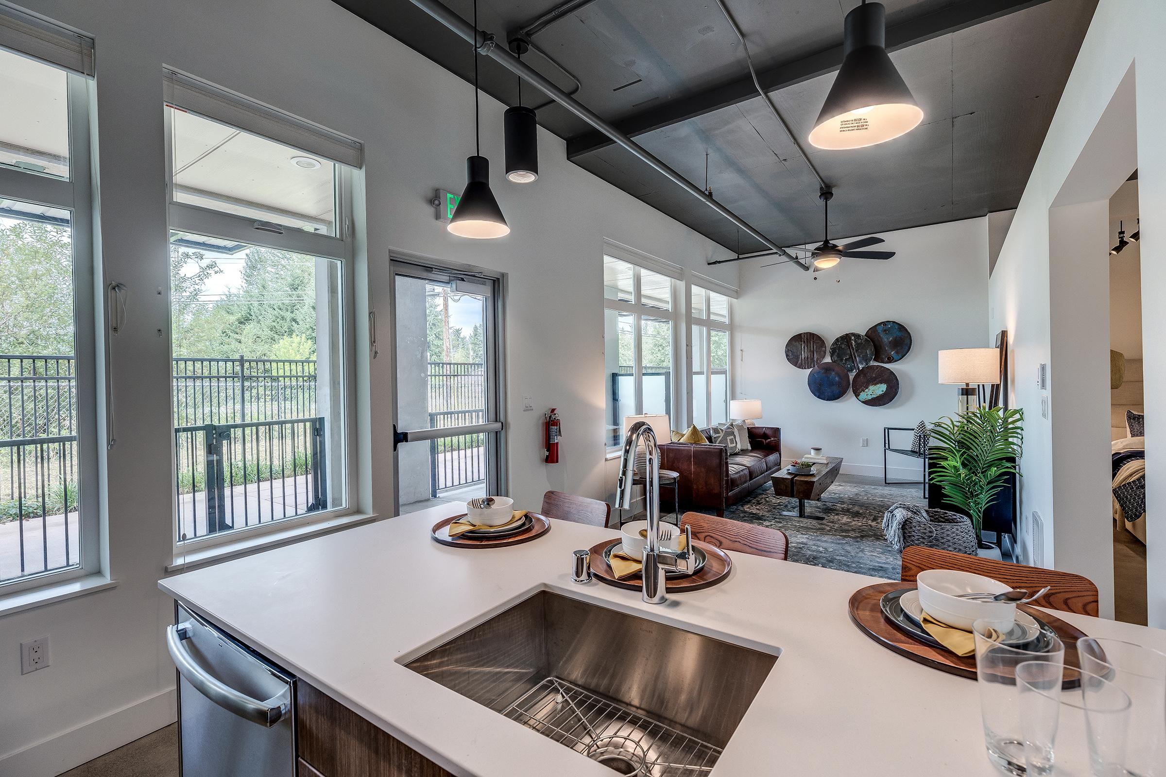 Modern apartment interior featuring a kitchen sink with dishes, an open layout, and large windows letting in natural light. A cozy living area is visible with comfortable seating, decorative wall art, and contemporary lighting fixtures. The space has a neutral color palette and stylish furnishings.