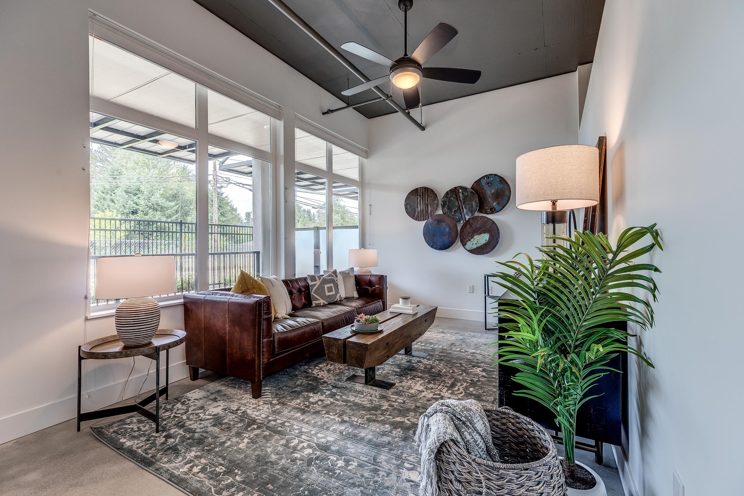 A modern living room featuring a brown leather sofa, a wooden coffee table, and decorative pillows. Large windows allow natural light, with a ceiling fan above. A floor lamp and a plant add warmth, while abstract wall art is displayed on the adjacent wall. The overall design is contemporary and inviting.