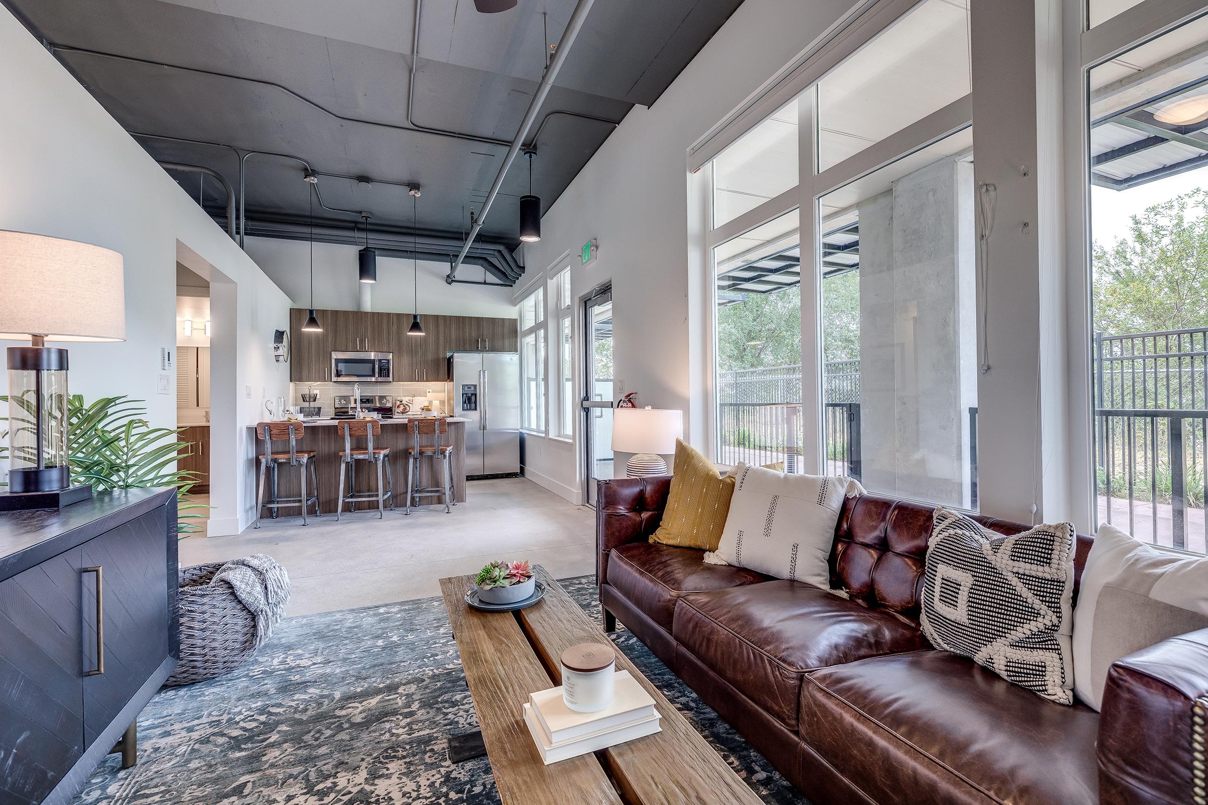 A modern living room featuring a brown leather sofa with decorative pillows, a wooden coffee table, and a stylish floor lamp. In the background, there's a dining area with sleek bar stools and a view of a kitchen. Large windows allow natural light to fill the space, complemented by contemporary decor elements.