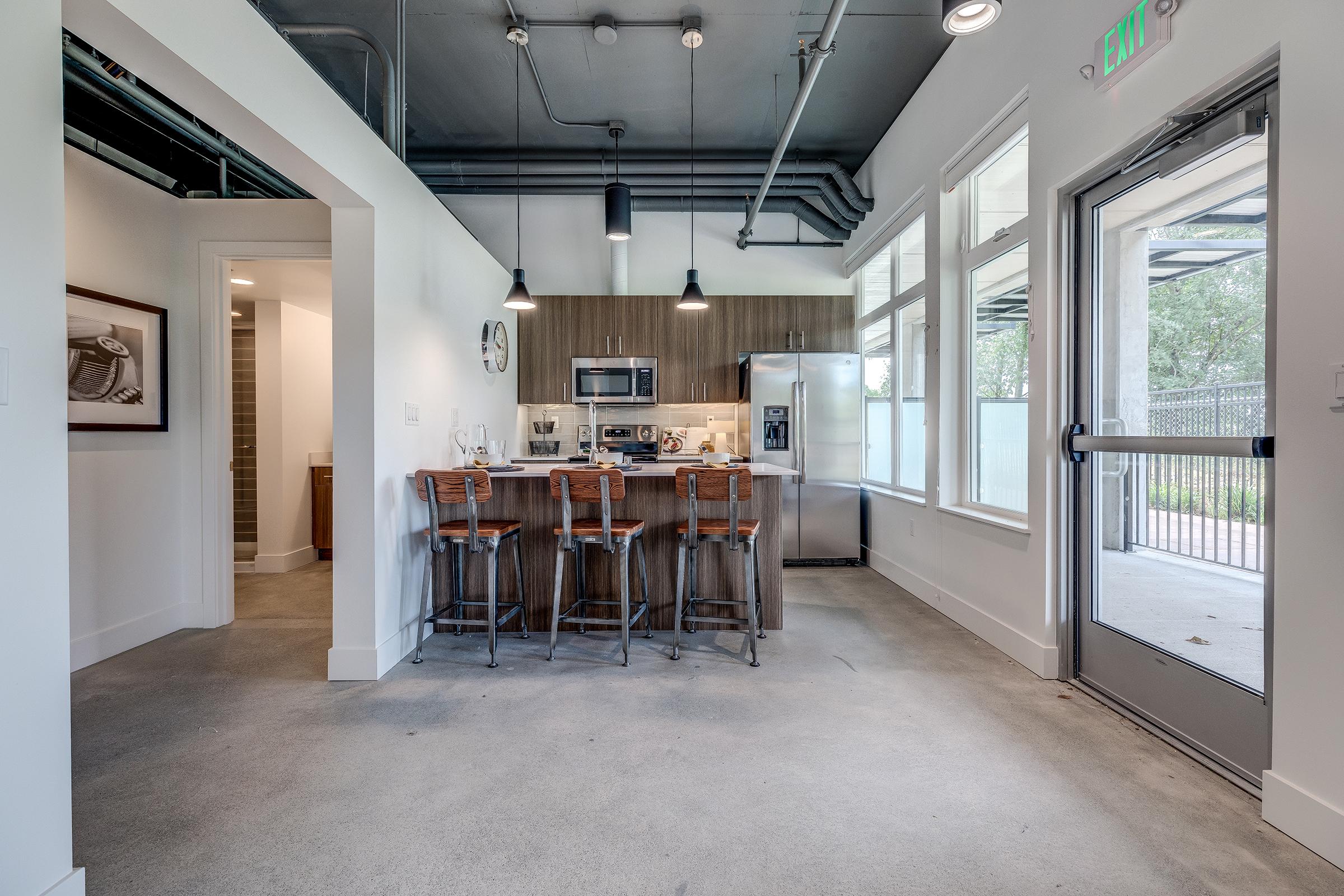 A modern kitchen with wooden cabinetry and stainless steel appliances. There are four wooden barstools at the kitchen island. Large windows allow natural light to fill the space, and the floor is smooth concrete. The room has a minimalist design with a neutral color palette.