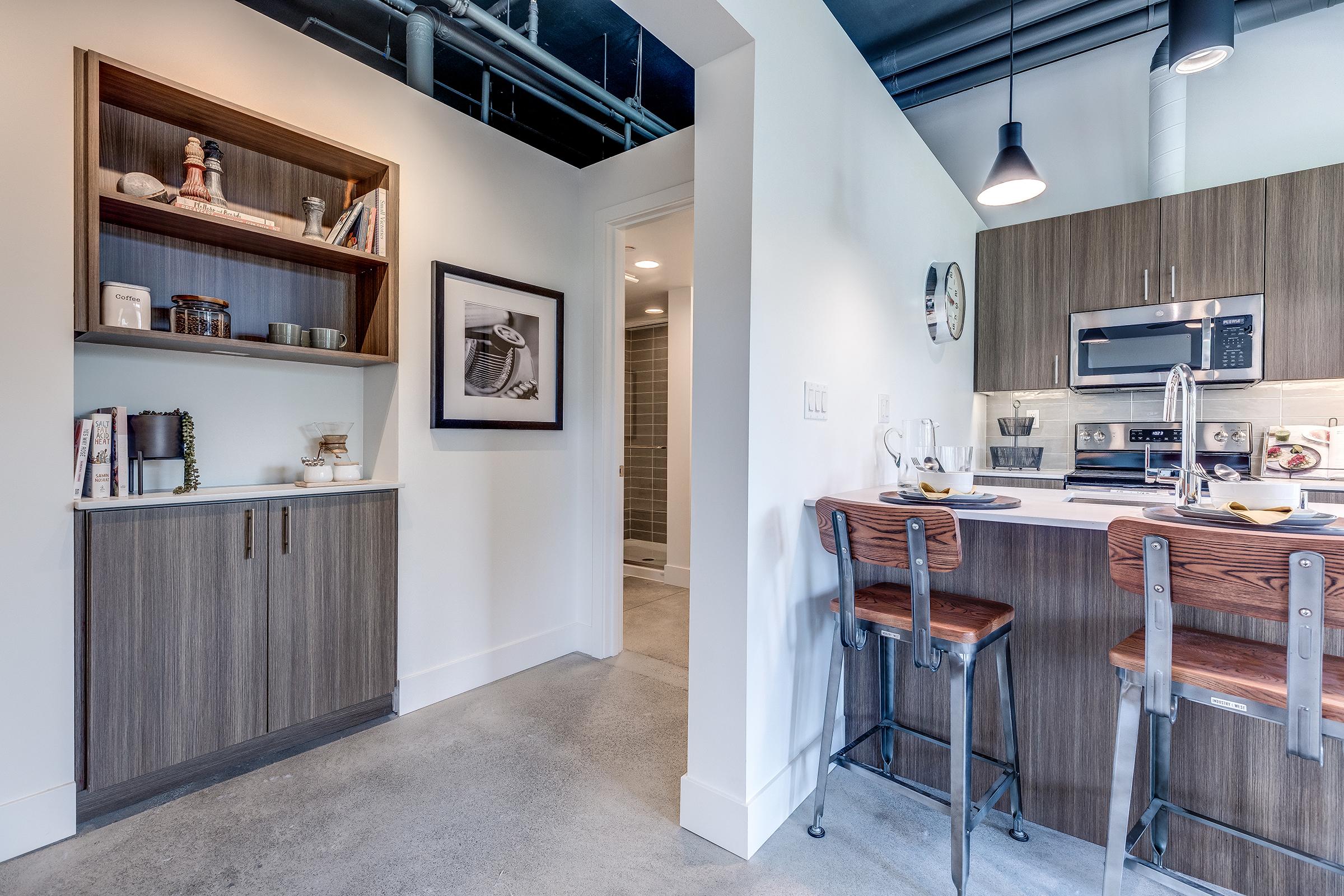 Modern kitchen and dining area featuring sleek wooden cabinetry, stainless steel appliances, and bar seating. The open layout includes a small shelf with decor items and a black-and-white framed artwork, complemented by industrial-style pendant lighting and polished concrete flooring.