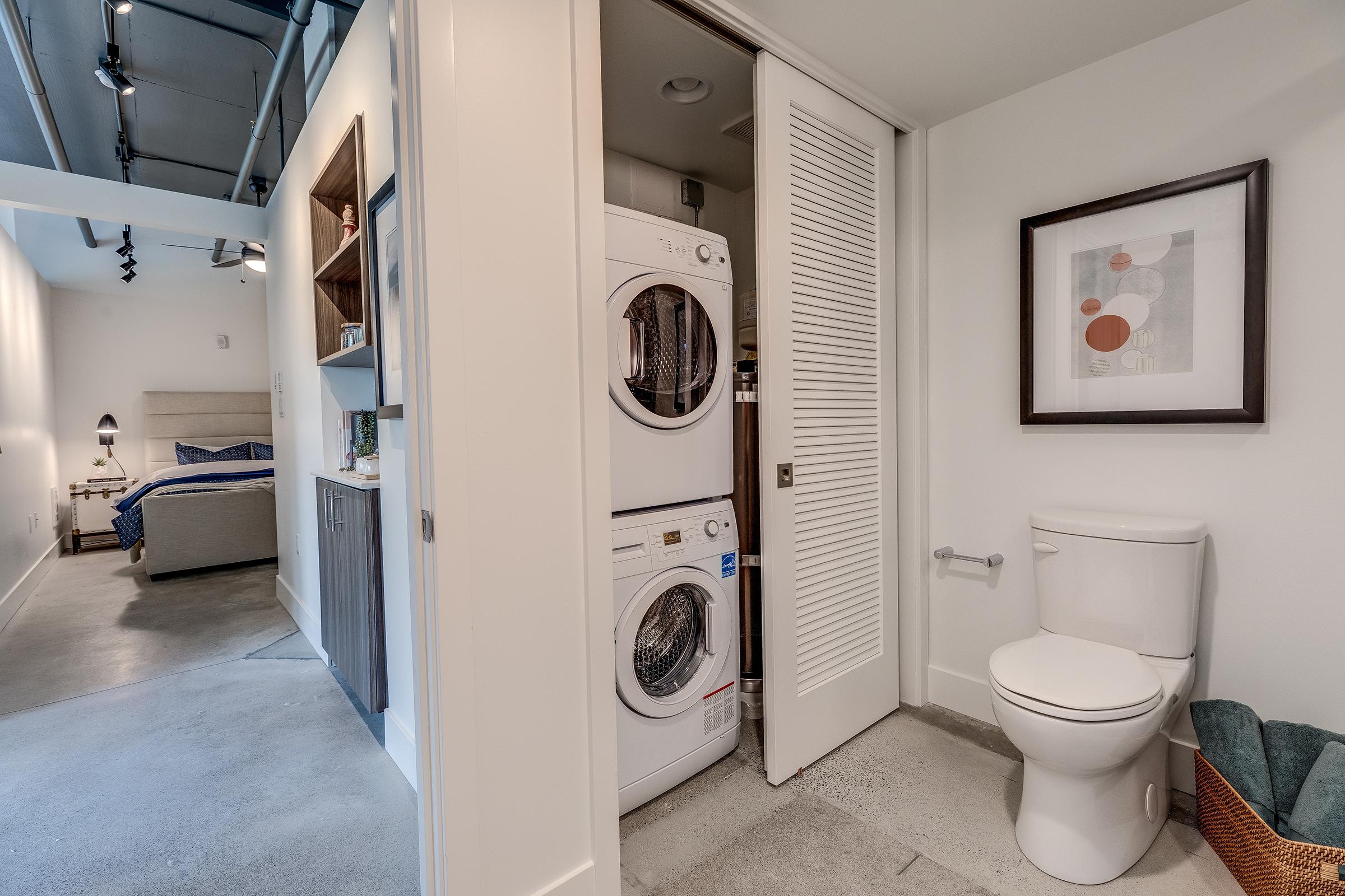 A compact laundry area featuring stacked washer and dryer units next to a small bathroom with a toilet. The space is modern with light-colored walls, and in the background, a bedroom is visible with a bed and decorative wall art. The flooring is concrete, adding to the contemporary aesthetic.