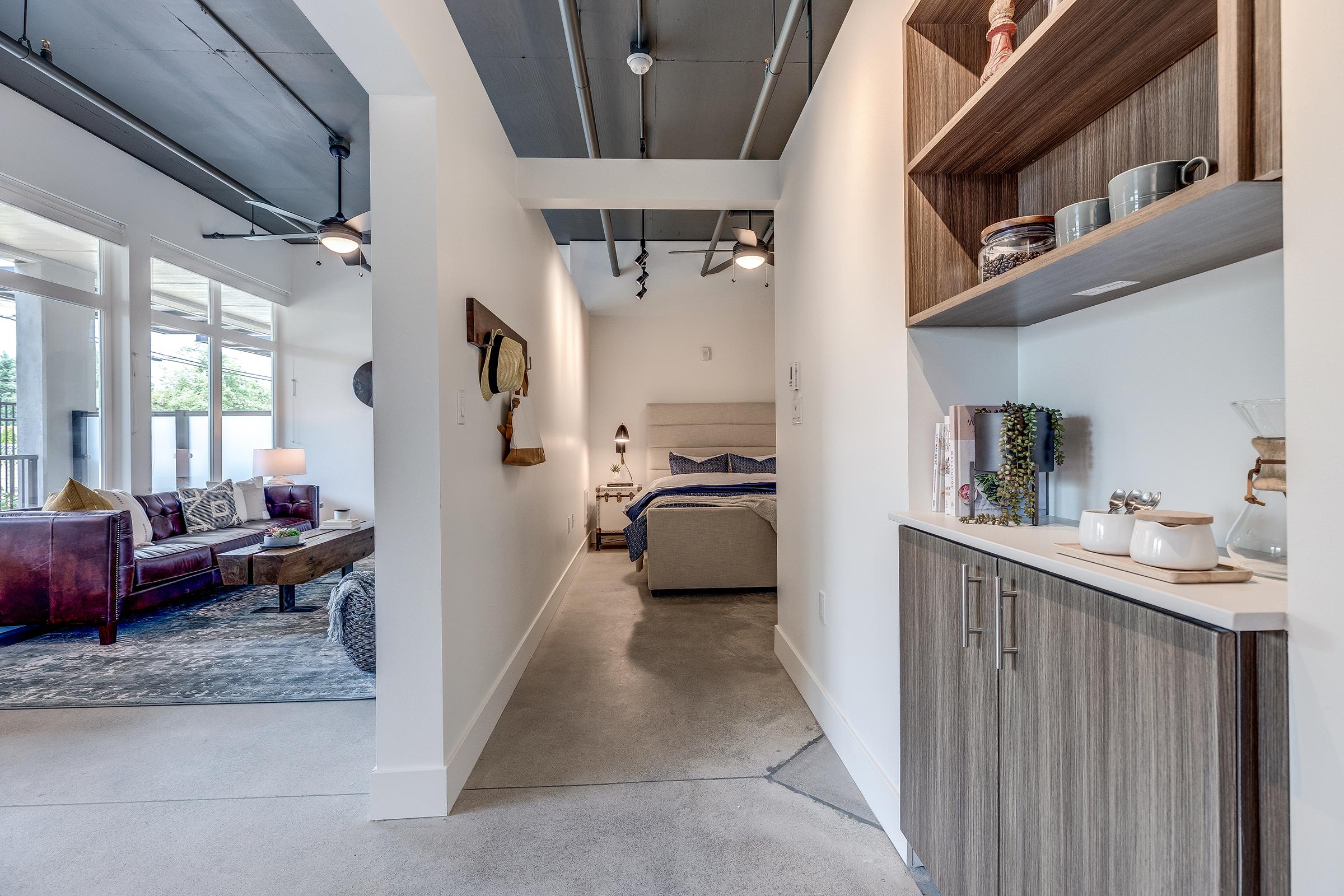A modern living space featuring an open layout. On the left, a cozy seating area with a sofa and decorative pillows, alongside a rug. To the right, a hallway leading to a bedroom with a bed and bedside tables. Wood cabinets and decorative items are visible, enhancing the stylish ambiance. Natural light from large windows brightens the area.