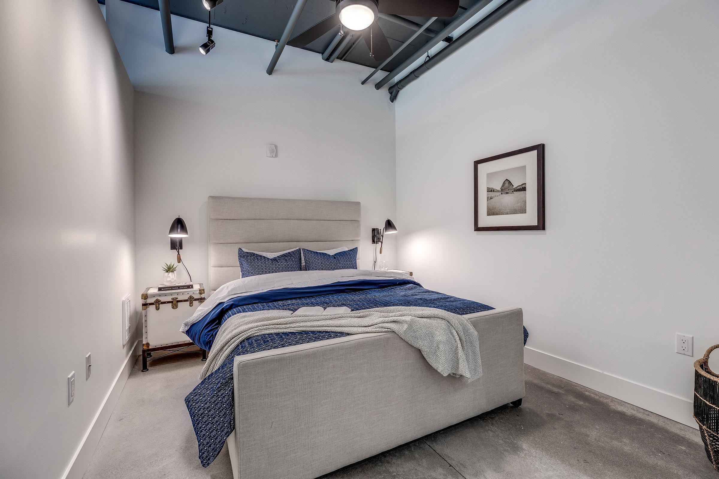 A cozy bedroom featuring a light gray upholstered bed with blue and gray bedding. The walls are white, and there's a framed black-and-white photo on the wall. Two modern bedside lamps provide soft lighting, and a small side table holds a decorative plant. Exposed ceiling beams and concrete floors add a modern touch.