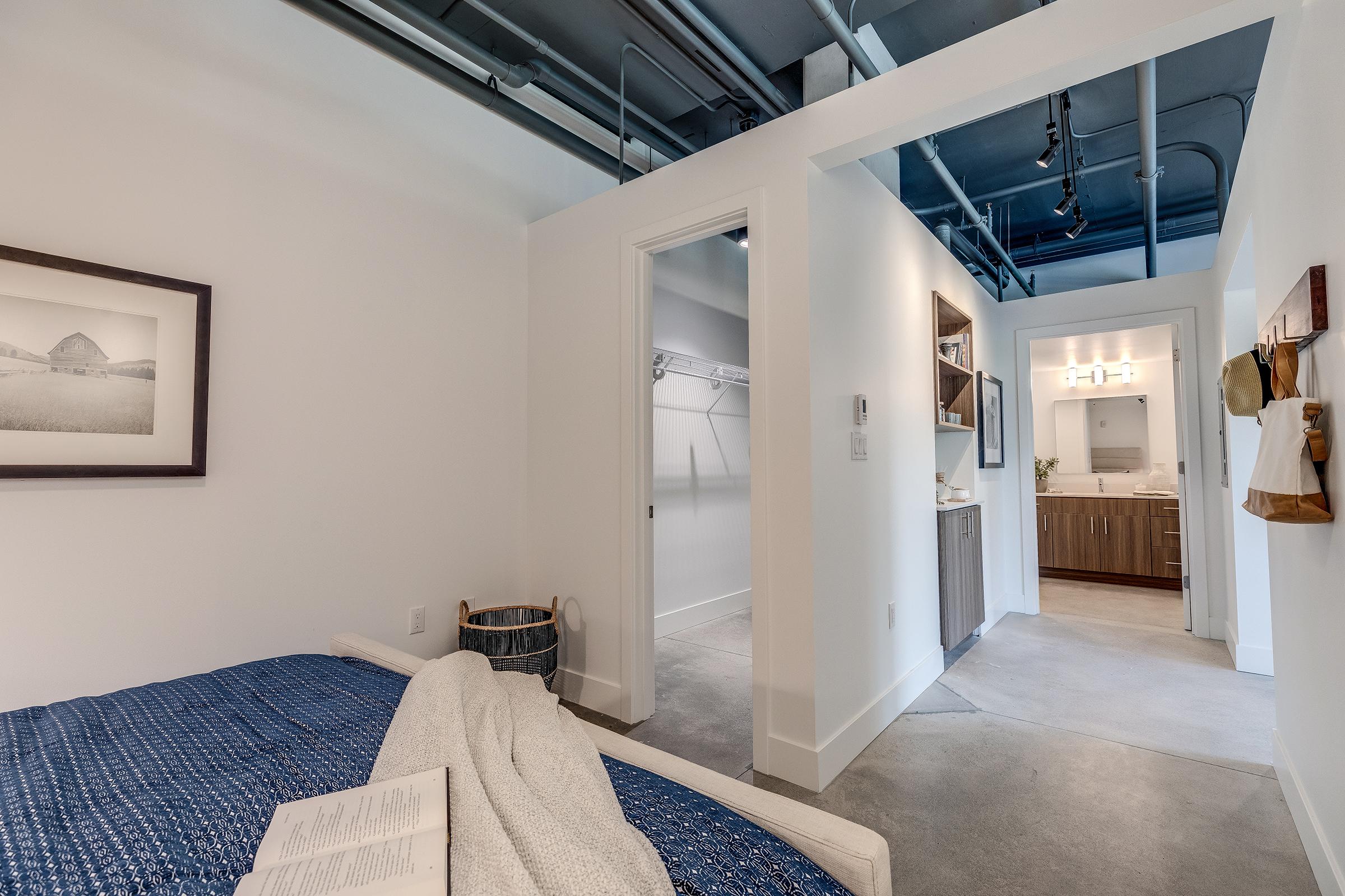 A modern bedroom featuring a blue quilt, with a closet space visible in the adjacent room. The area has clean lines, minimalistic decor, and soft lighting, creating a cozy and inviting atmosphere. The walls are painted white, and there is exposed piping on the ceiling.