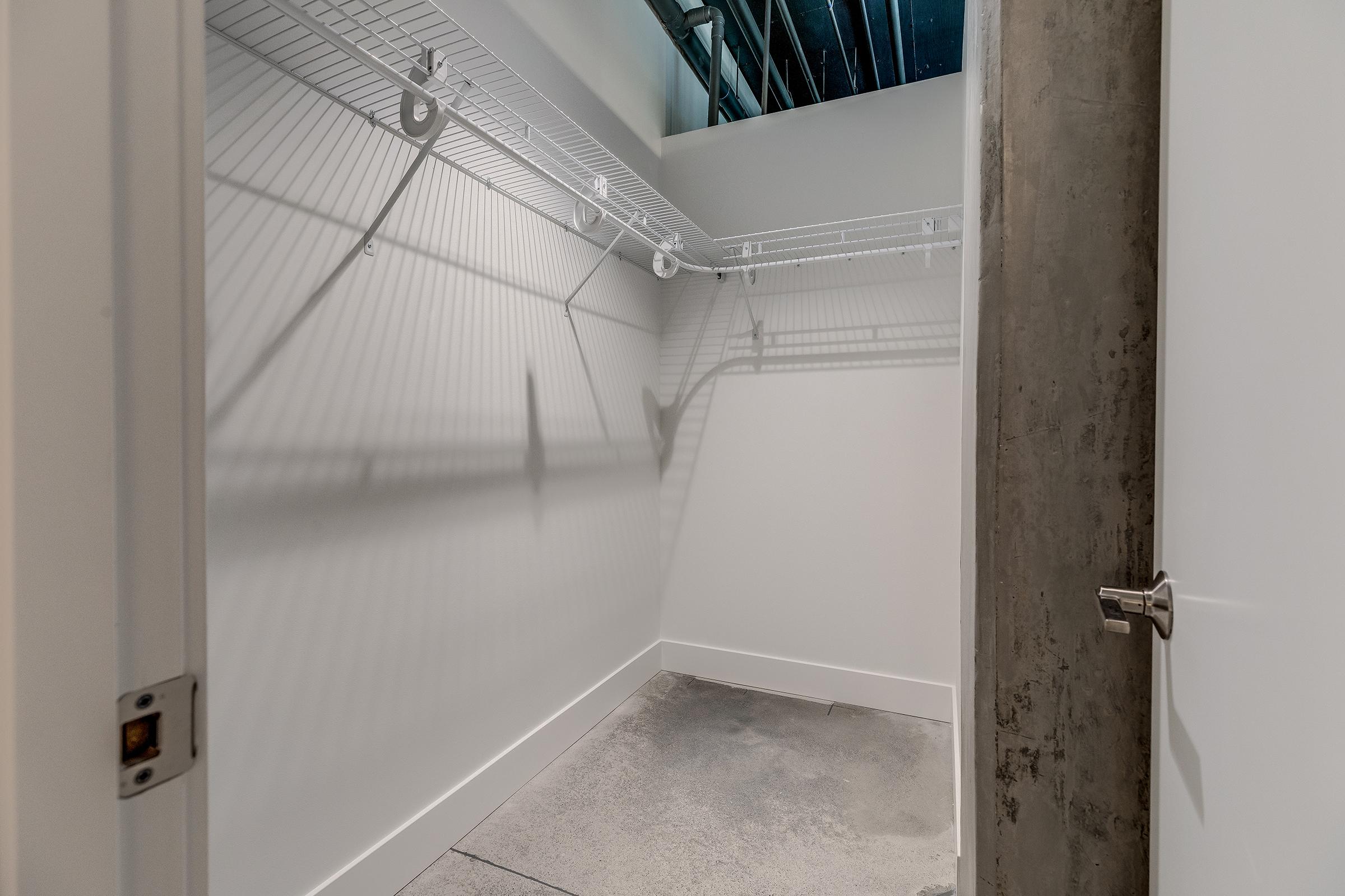 Empty walk-in closet featuring white wire shelving along one wall, a concrete floor, and a partially open door revealing a plain interior. The walls are painted a light color, and shadows from the shelves create patterns on the floor.
