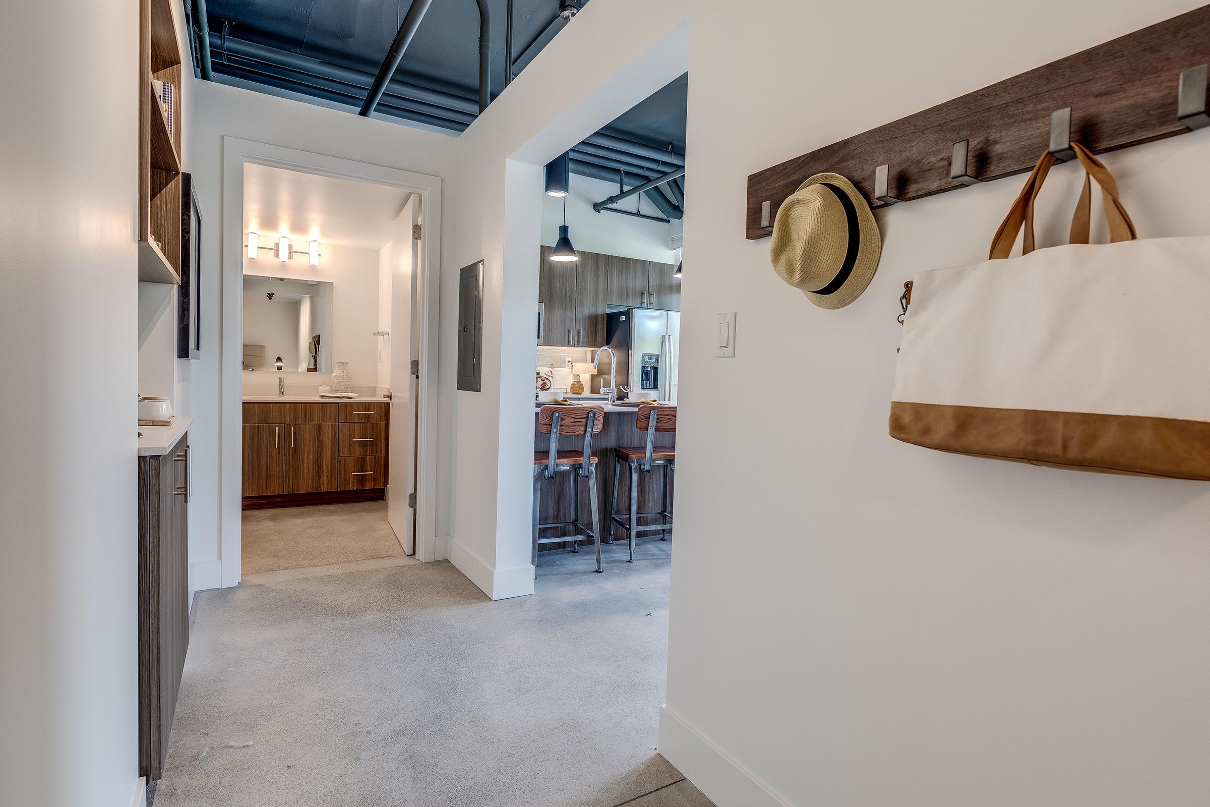 View of a modern hallway leading to a bathroom and kitchen area. The hallway features a light-colored wall, a wooden coat rack with hooks, a straw hat hanging, and a large canvas bag. The floor is polished concrete, adding to the contemporary design of the space.