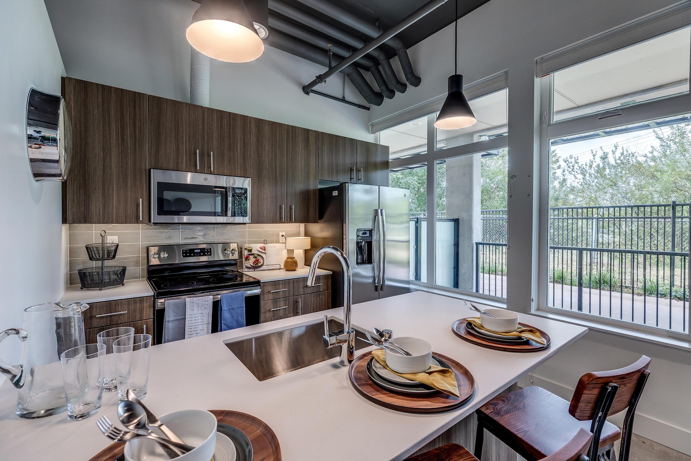 Lynwood, WA affordable apartments - Urban Center - Modern kitchen featuring stainless steel appliances, an island with plates, glassware, large windows, and pendant lights. Dark wood cabinetry just below exposed piping.