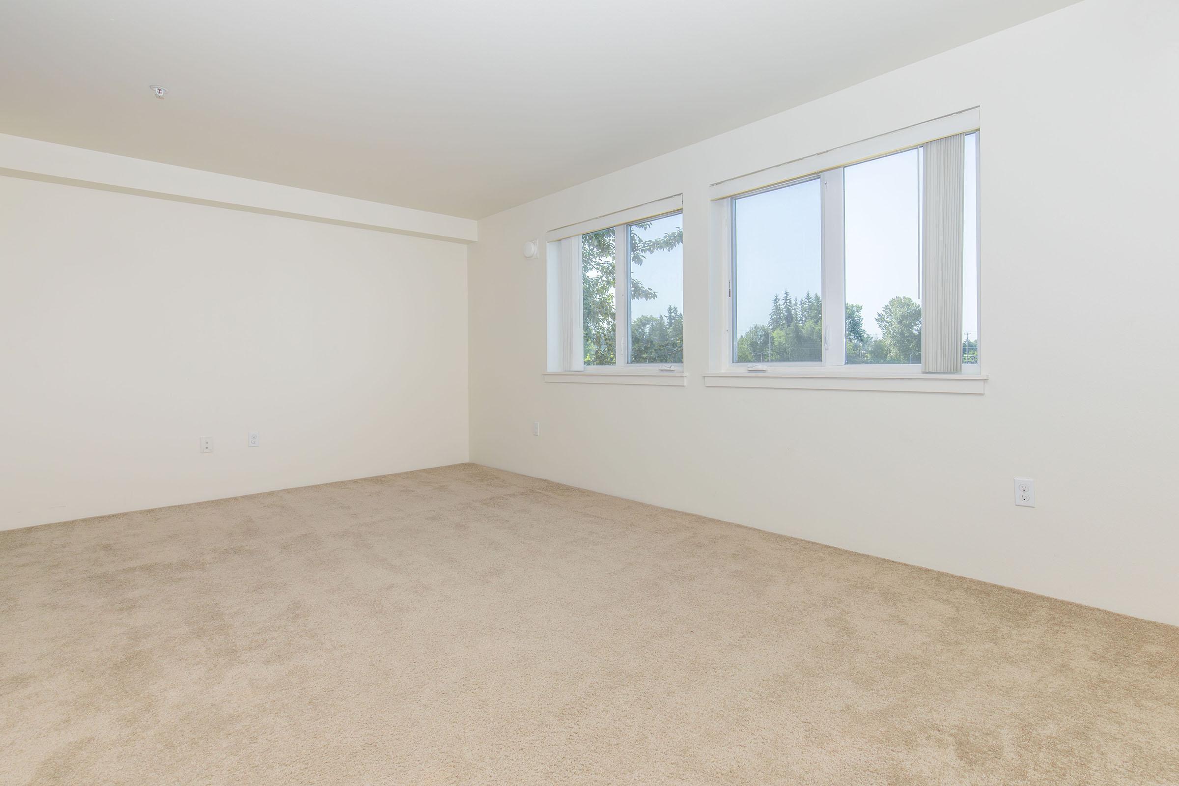 A spacious, empty room with beige carpet flooring and three large windows letting in natural light. The walls are painted a light color, creating a bright and airy atmosphere. There are no furnishings, making it ideal for personalization and decoration.