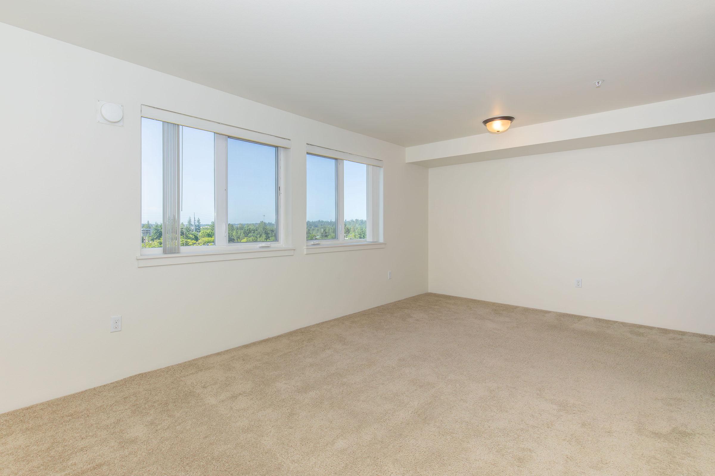 A spacious, empty room with light beige carpet and white walls. It features two large windows that let in natural light, offering a view of greenery outside. The ceiling is plain with a central light fixture. The room has a minimalist and bright atmosphere, suitable for various design possibilities.