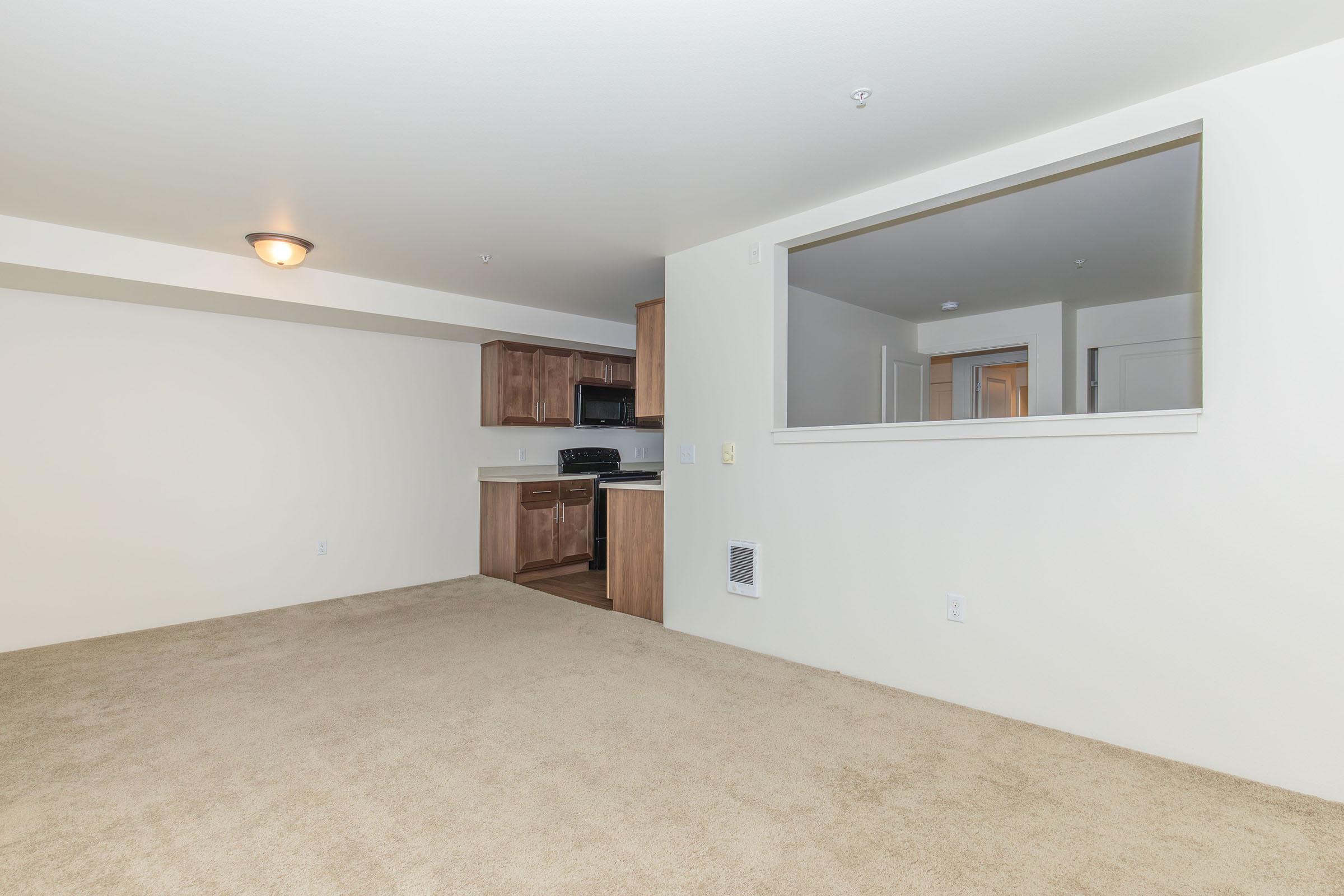 A spacious, empty room with light-colored walls and carpet. There is a kitchen area with wooden cabinets, a stove, and a microwave visible through an opening. The layout is open, and there is a ceiling light fixture. The overall ambiance is clean and ready for furnishing.