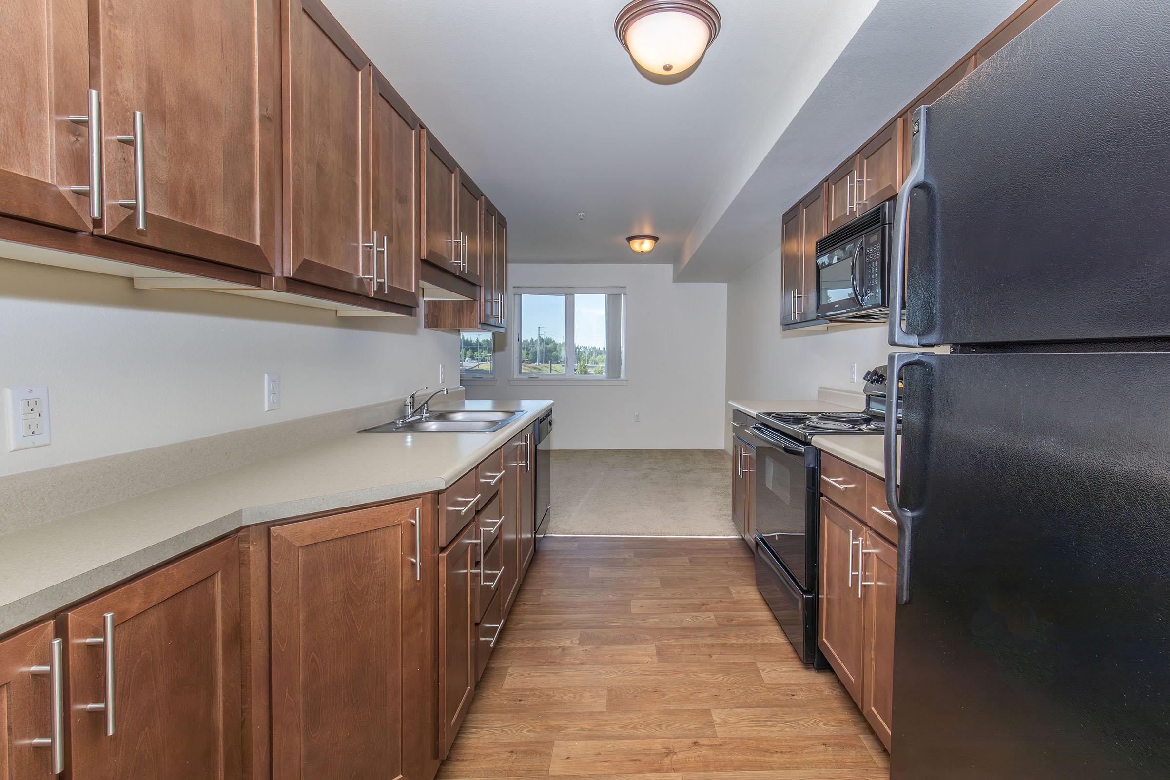 A spacious kitchen featuring dark wood cabinets, a black refrigerator, and a black stove. The countertops are light-colored, and there are dual sinks. Natural light comes from a nearby window, providing a view of the outdoors. The flooring is a warm brown, complementing the kitchen's overall design.