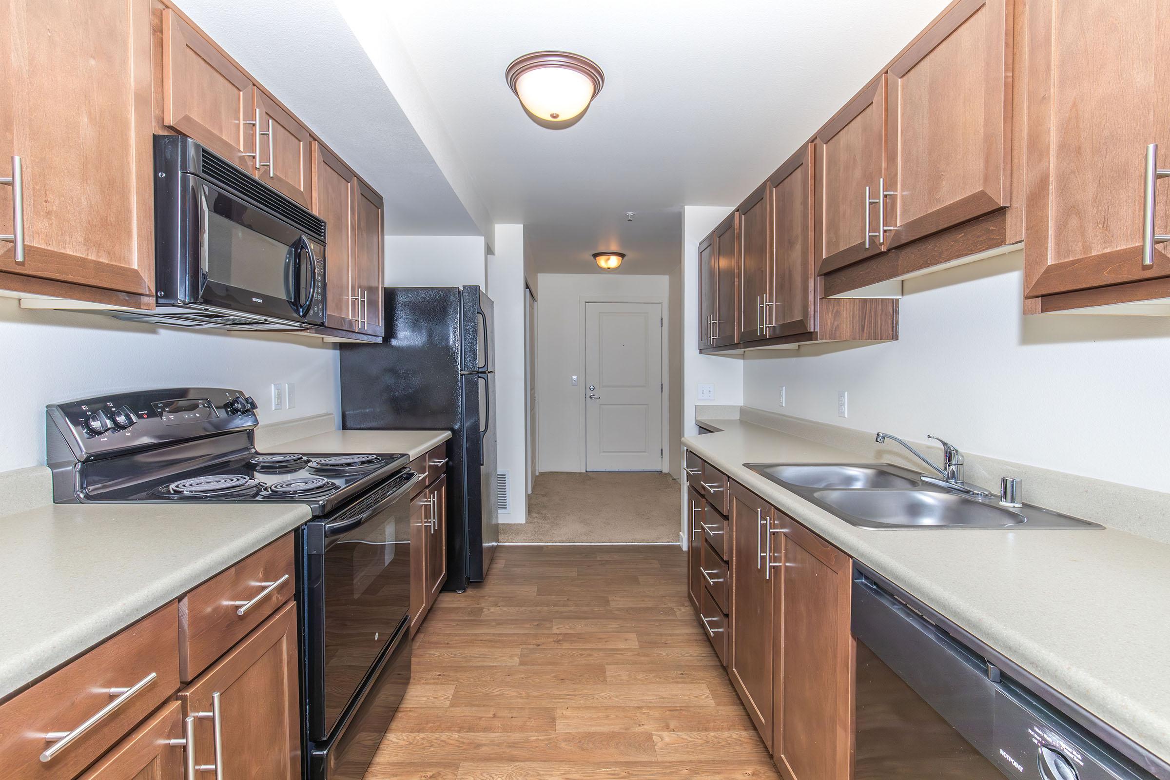 Modern kitchen featuring dark wood cabinets, stainless steel appliances including a microwave and oven, and a double sink. The countertop is light-colored, and the floor is a warm wood tone. There is an entrance visible at the far end of the room, along with a black refrigerator.