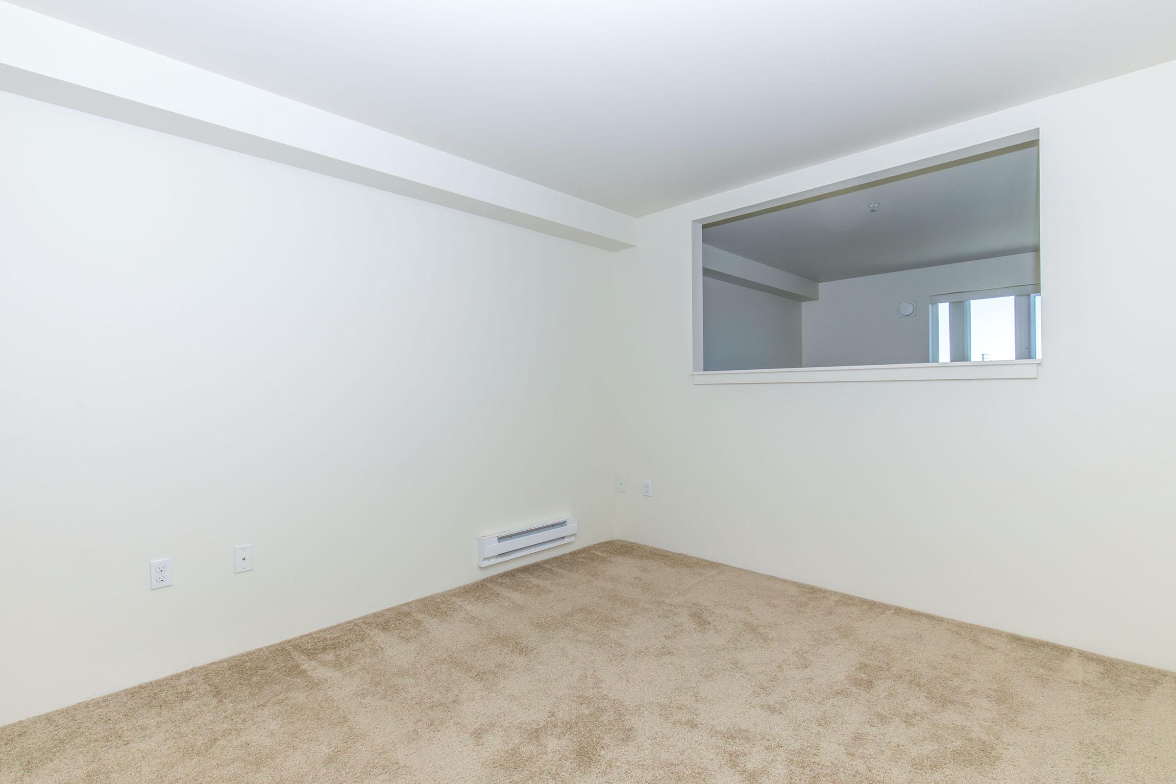 A sparsely furnished room with light beige carpet and white walls. There is a large window with a mirror above it, allowing natural light to enter. The space appears empty and clean, suitable for personalization or decoration.