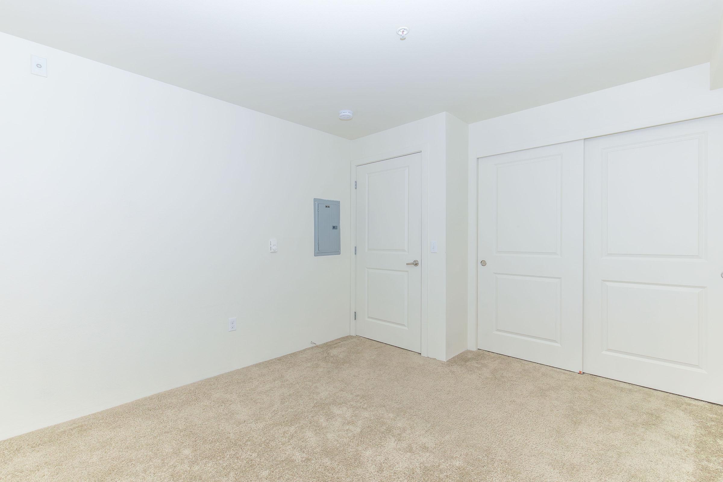 An empty room with light beige carpet, featuring white walls and a white door. There is a small electrical panel mounted on the wall, and a closet with double doors on one side. The space is well-lit and uncluttered, creating a minimalist aesthetic.