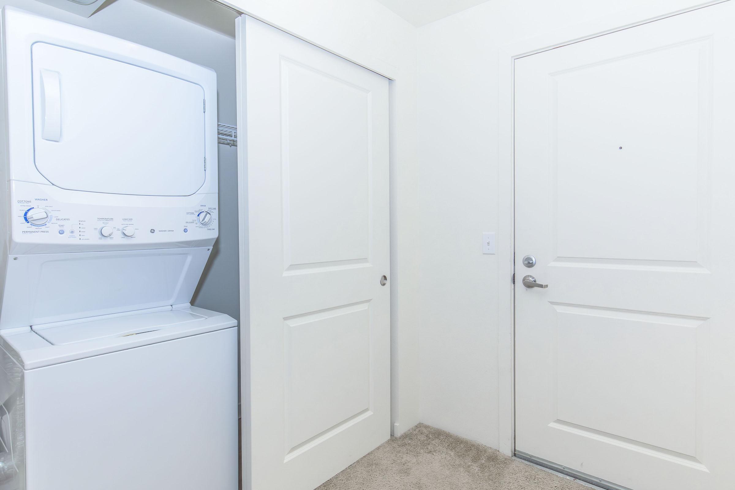 A laundry area featuring a stacked washer and dryer unit next to a closed door. The walls are painted white, and there is carpet flooring. A door leading to the outside or another room is visible, with a small peephole. The overall space appears clean and well-maintained.