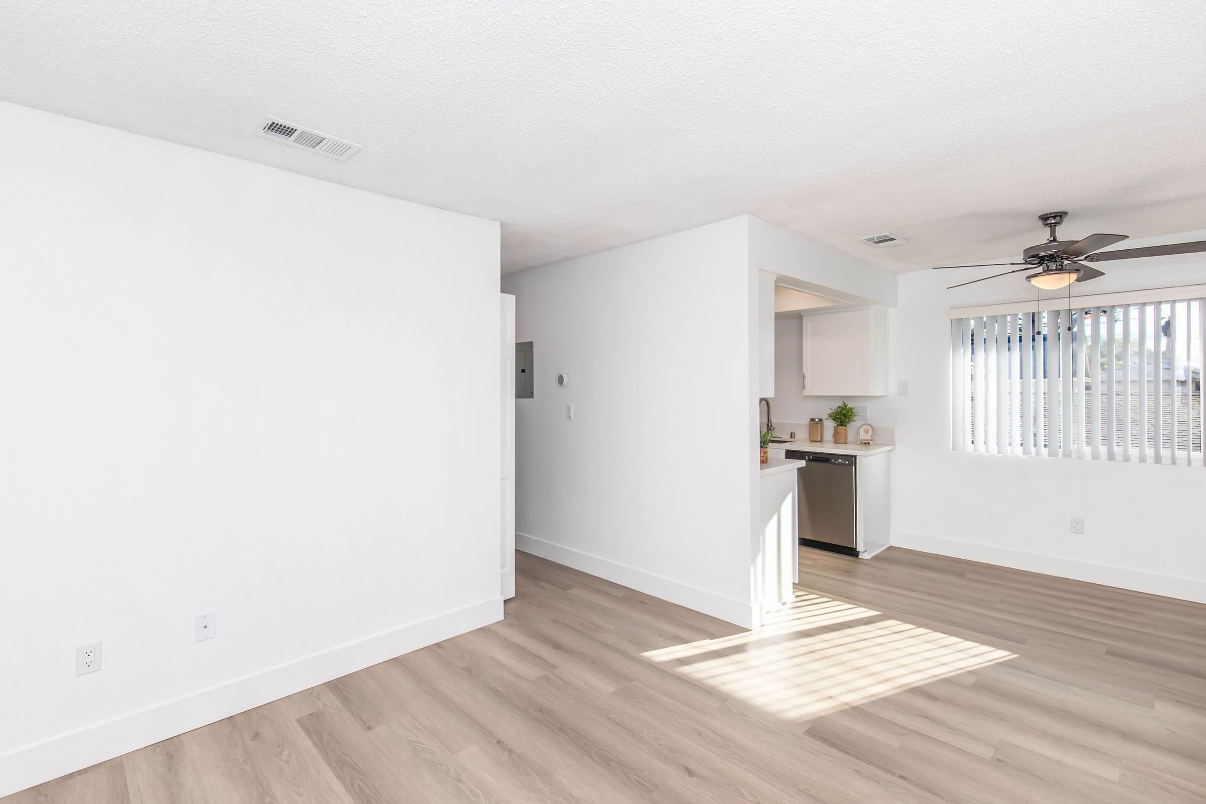 A bright, modern living space with light-colored walls and wooden flooring. The room features a ceiling fan, a wall-mounted air conditioning unit, and a doorway leading to a kitchen area with appliances. Large blinds cover the window, allowing natural light to flood the room.