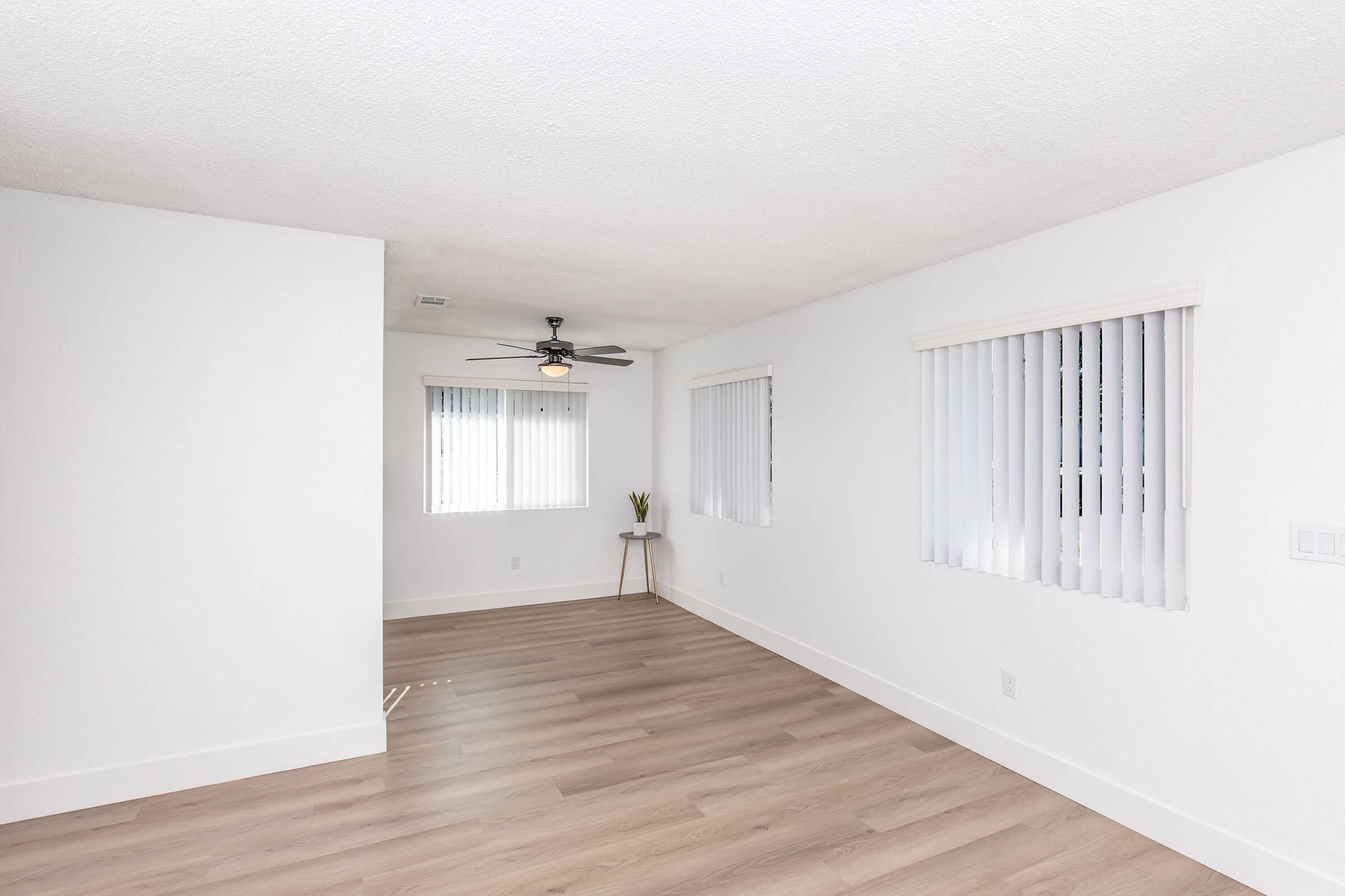 A well-lit interior room featuring light-colored walls and laminate flooring. It has a ceiling fan, two vertical-blind windows allowing natural light, and a small table with a plant in the corner. The space is simple and modern, promoting an airy and spacious atmosphere.