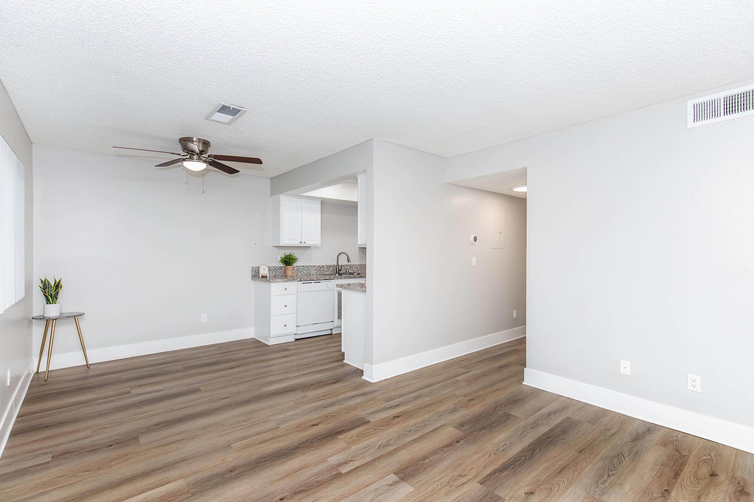 Bright, modern living space with light gray walls and wood-style flooring. Features include a ceiling fan, kitchen area visible in the background with white cabinetry and a granite countertop. A small plant on a side table adds a touch of greenery. Natural light coming through a window enhances the airy feel.