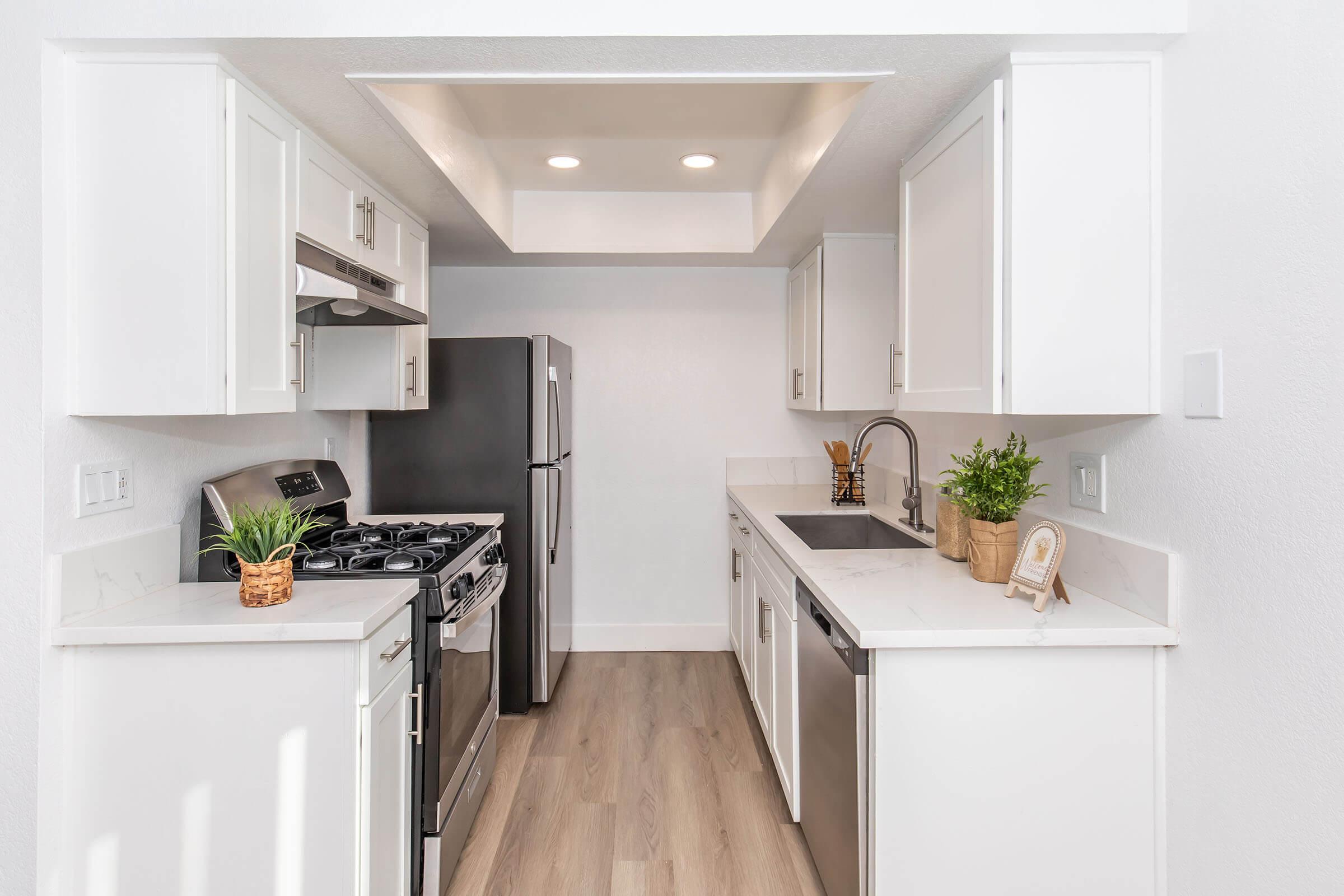 A modern kitchen featuring white cabinetry, stainless steel appliances, and stone countertops. The layout includes a gas stove, refrigerator, sink, and decorative plants, with a bright, airy feel created by natural light and neutral colors.