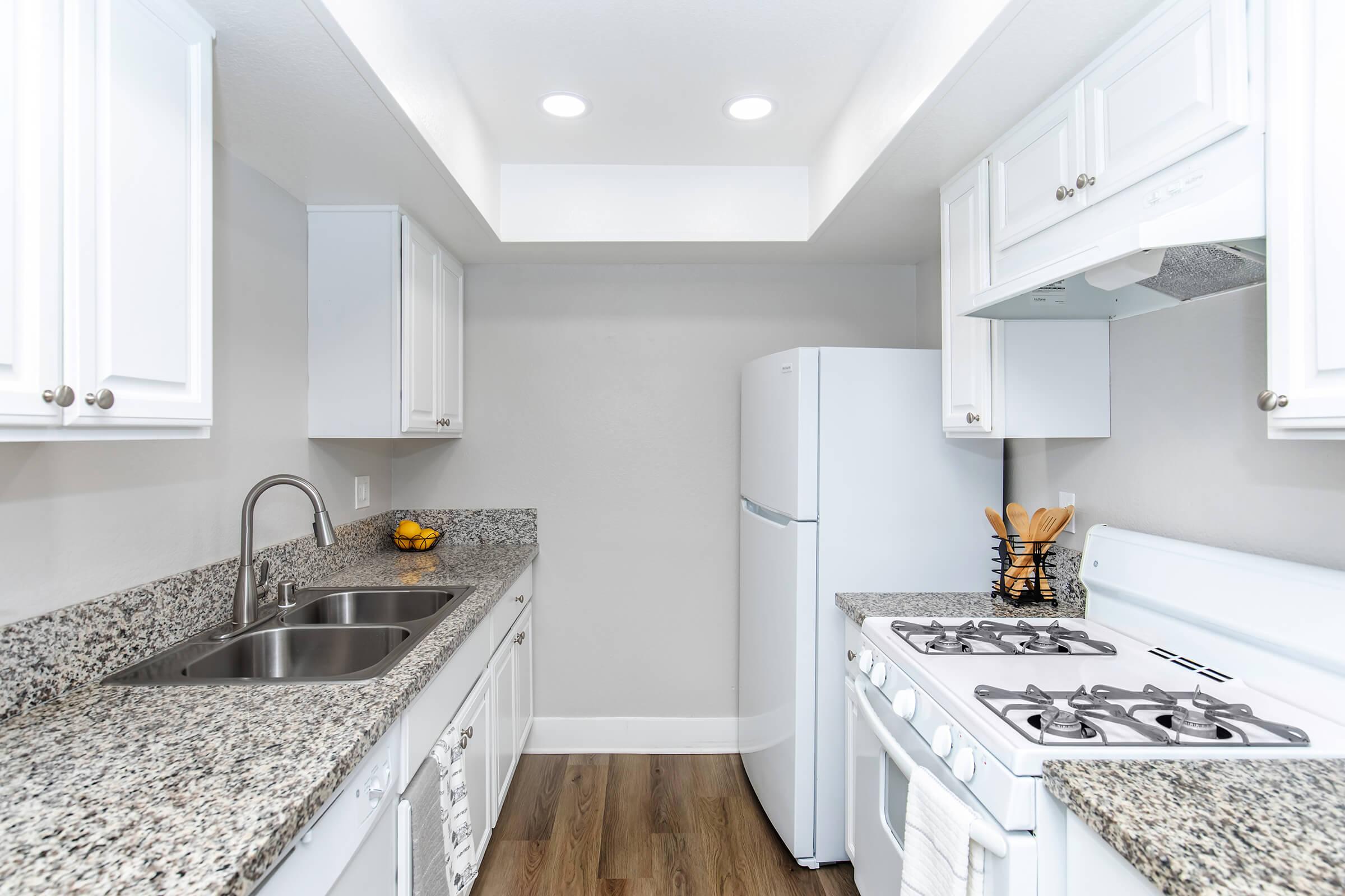 Modern kitchen featuring white cabinetry, a granite countertop, stainless steel sink, and a gas stove. A white refrigerator is visible, along with a bowl of fruits on the counter. The space is well-lit with recessed lighting and has a clean, contemporary design.