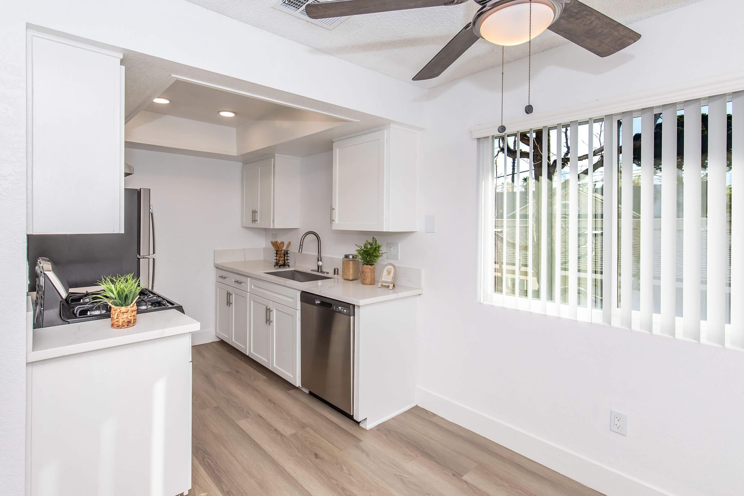 A modern kitchen featuring white cabinetry, stainless steel appliances, and a marble countertop. The space includes a gas stove, a dishwasher, and a sink. Natural light enters through a window with horizontal blinds, complementing the light wood flooring and sleek design.