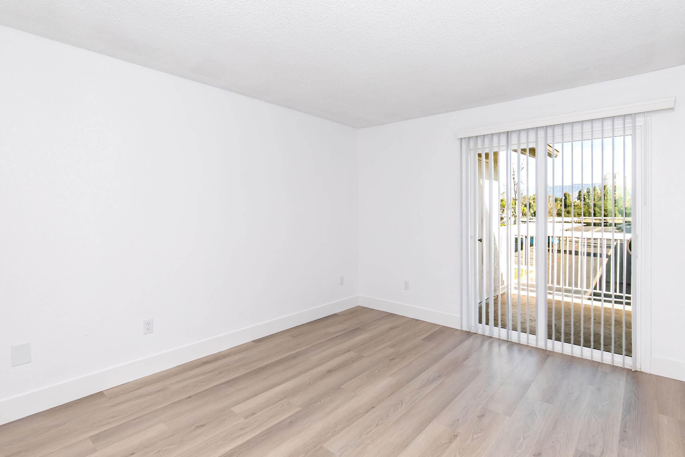 A bright, empty room with white walls and light wood flooring. Sliding glass doors lead to a small outdoor space with a view of greenery. The room has natural light and a minimalist design, creating a spacious feel.