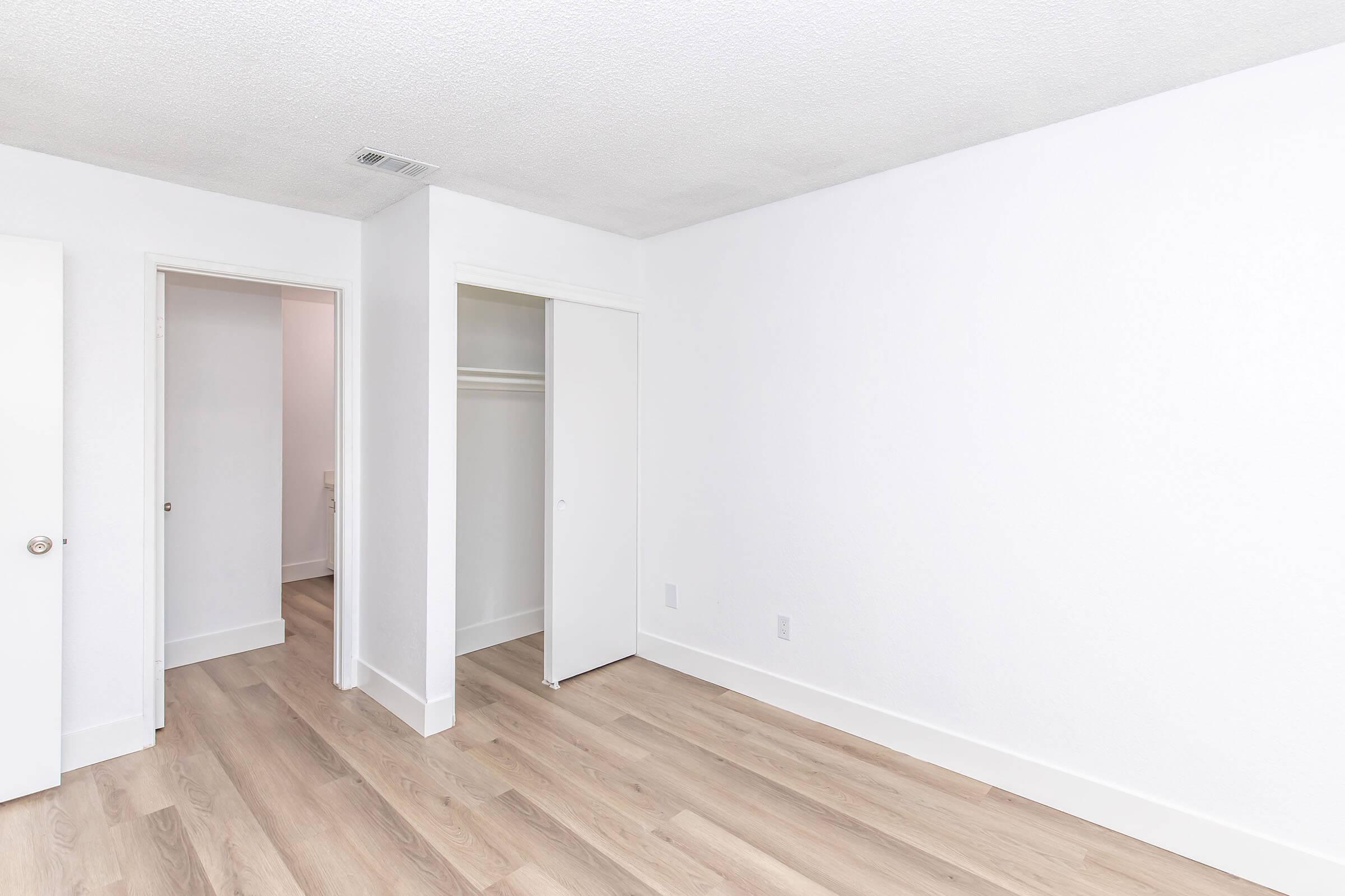 Empty room with white walls and a light wooden floor. One side features a closet with sliding doors, while there are two doorways leading to adjacent spaces. The overall atmosphere is bright and minimalistic, creating a spacious feel.