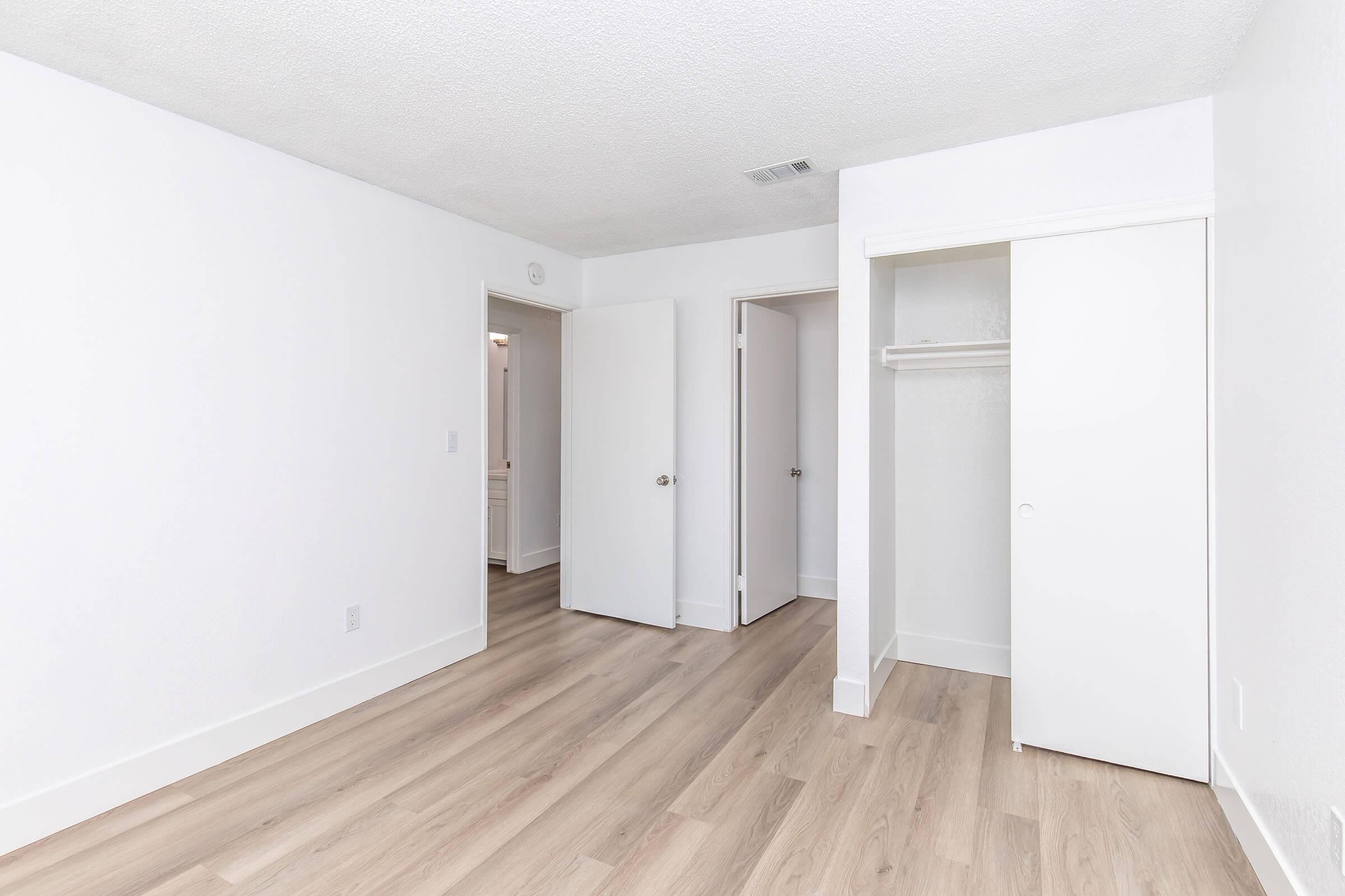 A bright, empty room featuring light-colored walls and a sleek, wooden floor. One wall has a closet with sliding doors, and there are two doors leading to other spaces. The overall atmosphere is clean and minimalist, suitable for various interior design options.