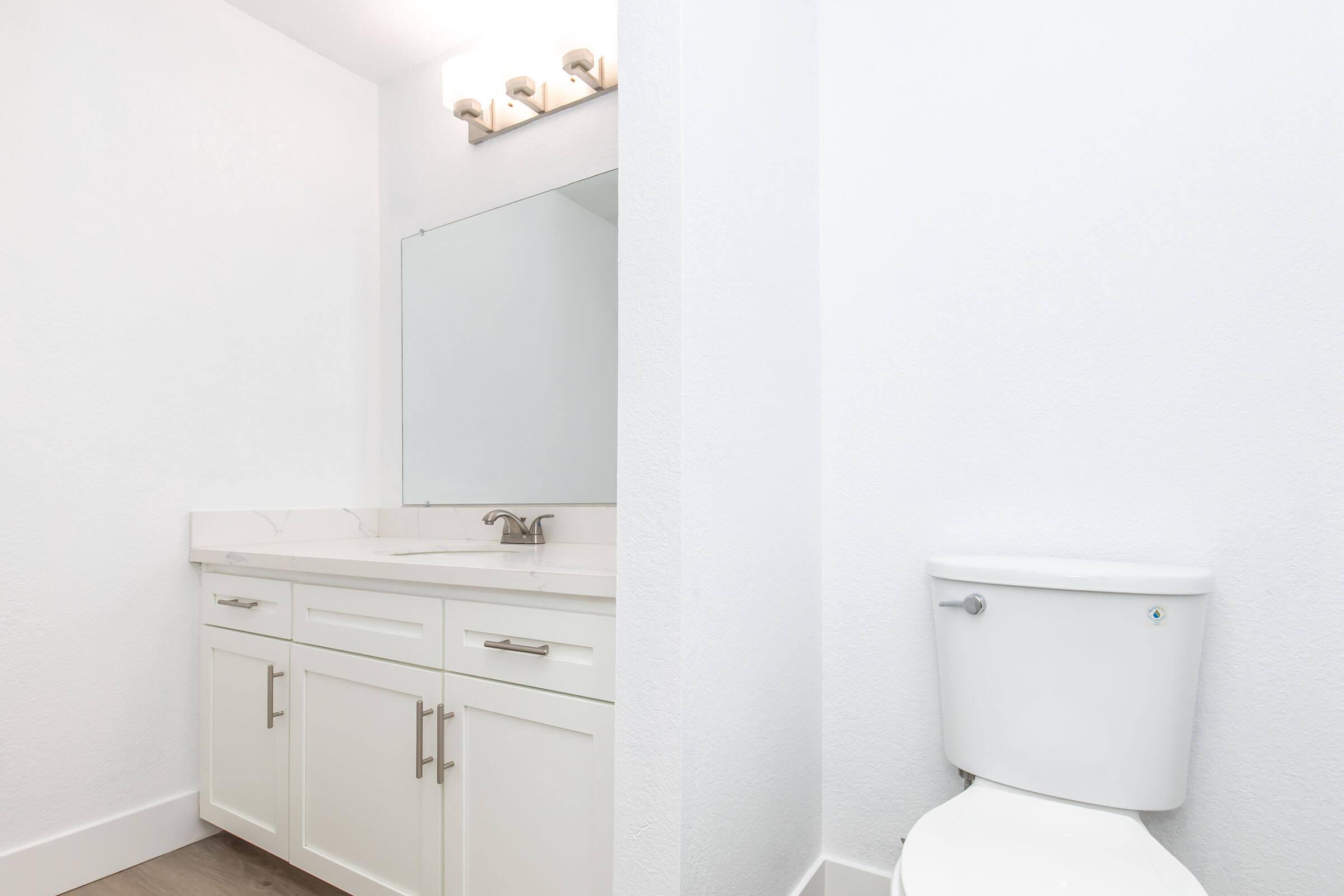 A clean and modern bathroom featuring a white vanity with a marble countertop, a large mirror, and a wall-mounted light fixture. To the right, there is a white toilet, and the walls are painted a bright white, creating a fresh and spacious feel. The flooring is light-colored, adding to the overall brightness.