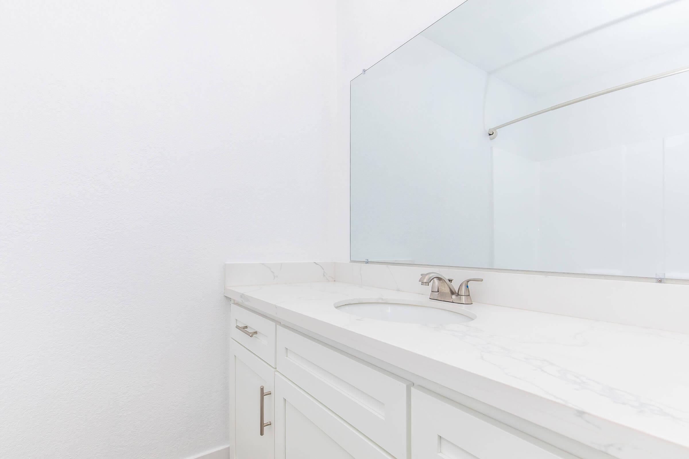 A clean, modern bathroom with a white marble countertop and a rectangular mirror. The sink has a stylish faucet. The walls are painted white, creating a bright, minimalist aesthetic. There's a towel rack above the sink, and the overall design emphasizes simplicity and elegance.