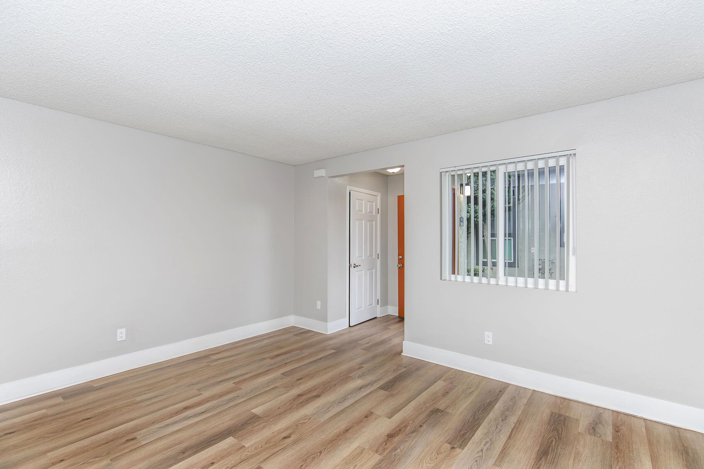 A brightly lit, recently renovated room with light gray walls and wooden flooring. The space includes a door leading to another room and a window with vertical blinds. The overall atmosphere is clean and minimalistic, making it suitable for various uses.