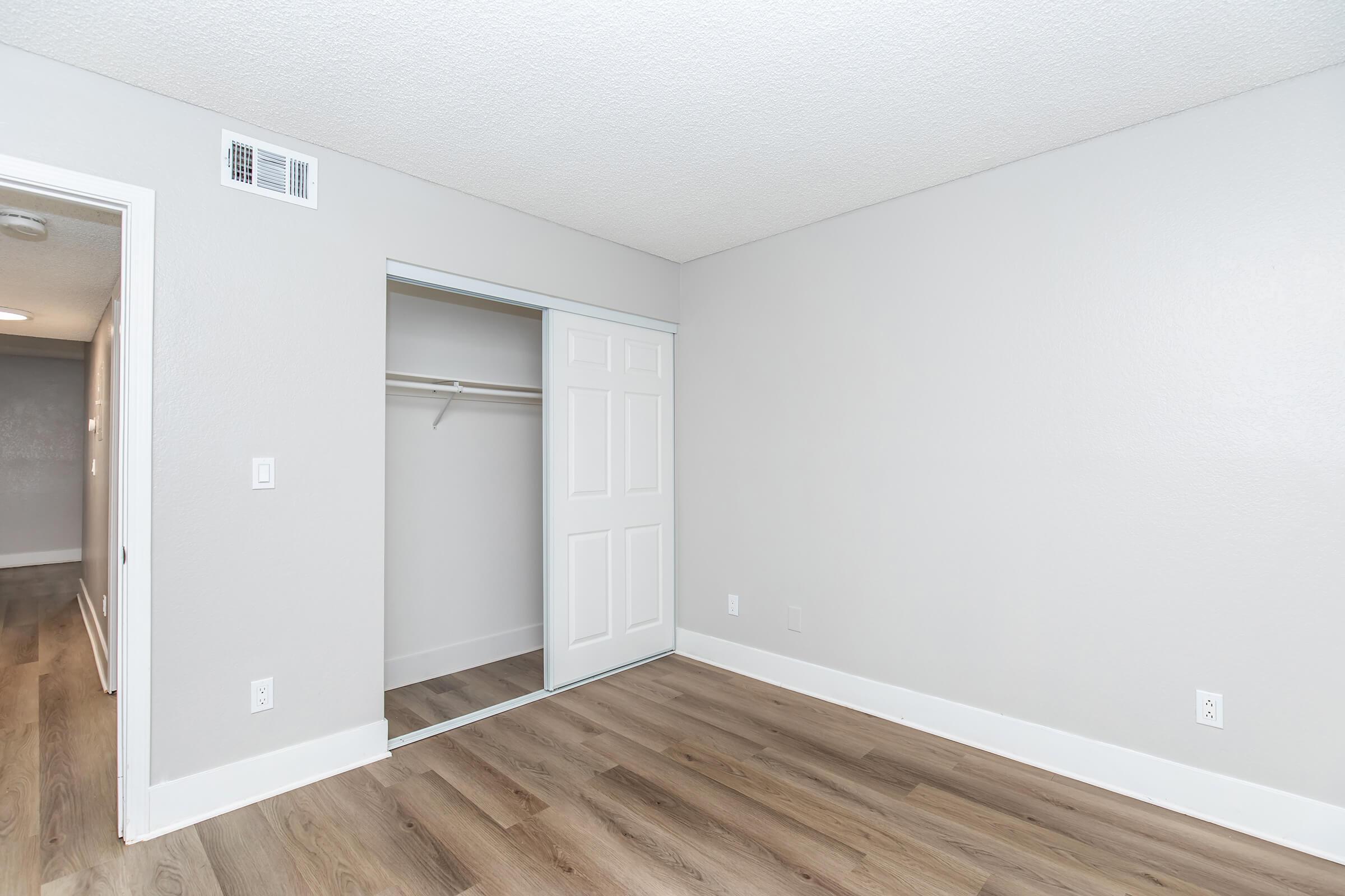Empty room with light gray walls and laminate flooring. There is a closet with a sliding white door on the left side, and no furniture or decorations are present. The lighting is bright, creating a spacious and clean atmosphere.