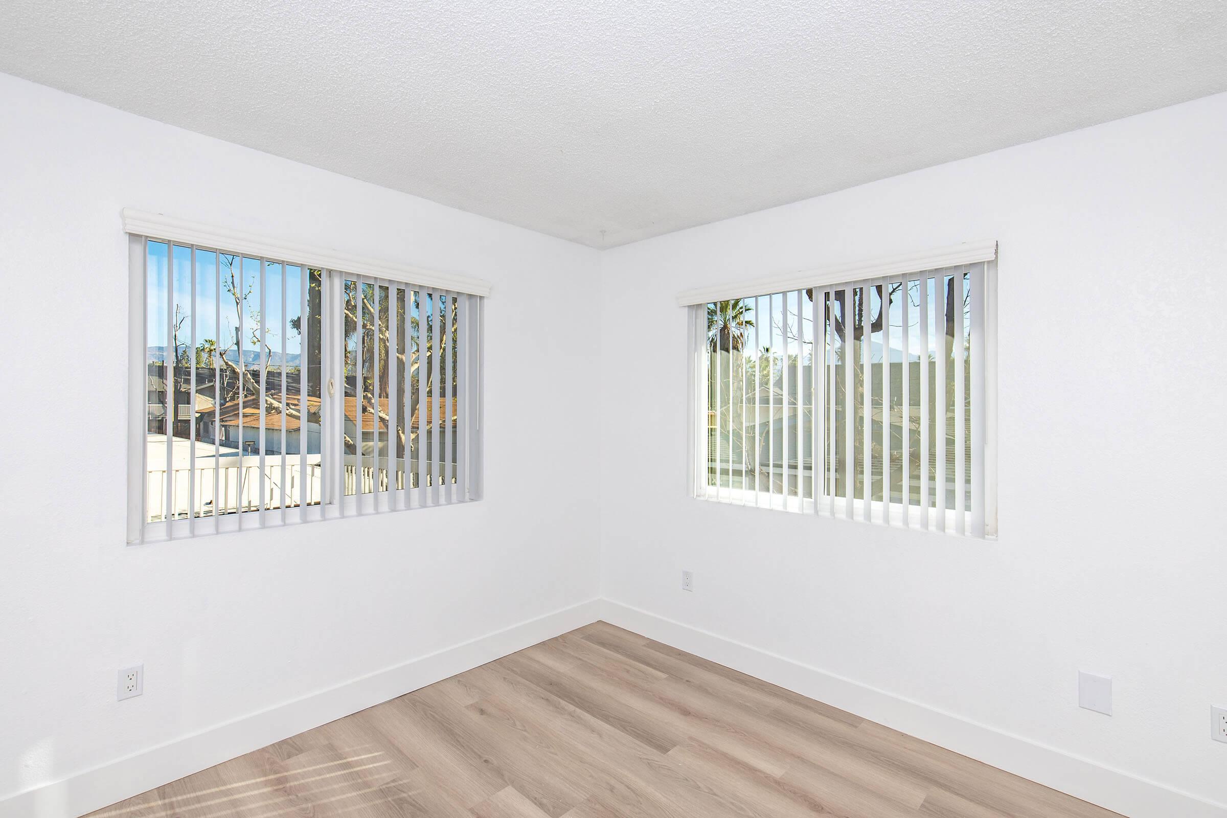 Empty room with white walls and light wood flooring. Two windows with vertical blinds allow natural light, and a view of trees outside is visible. The space is bright and minimalistic, suited for various layouts or furnishings.