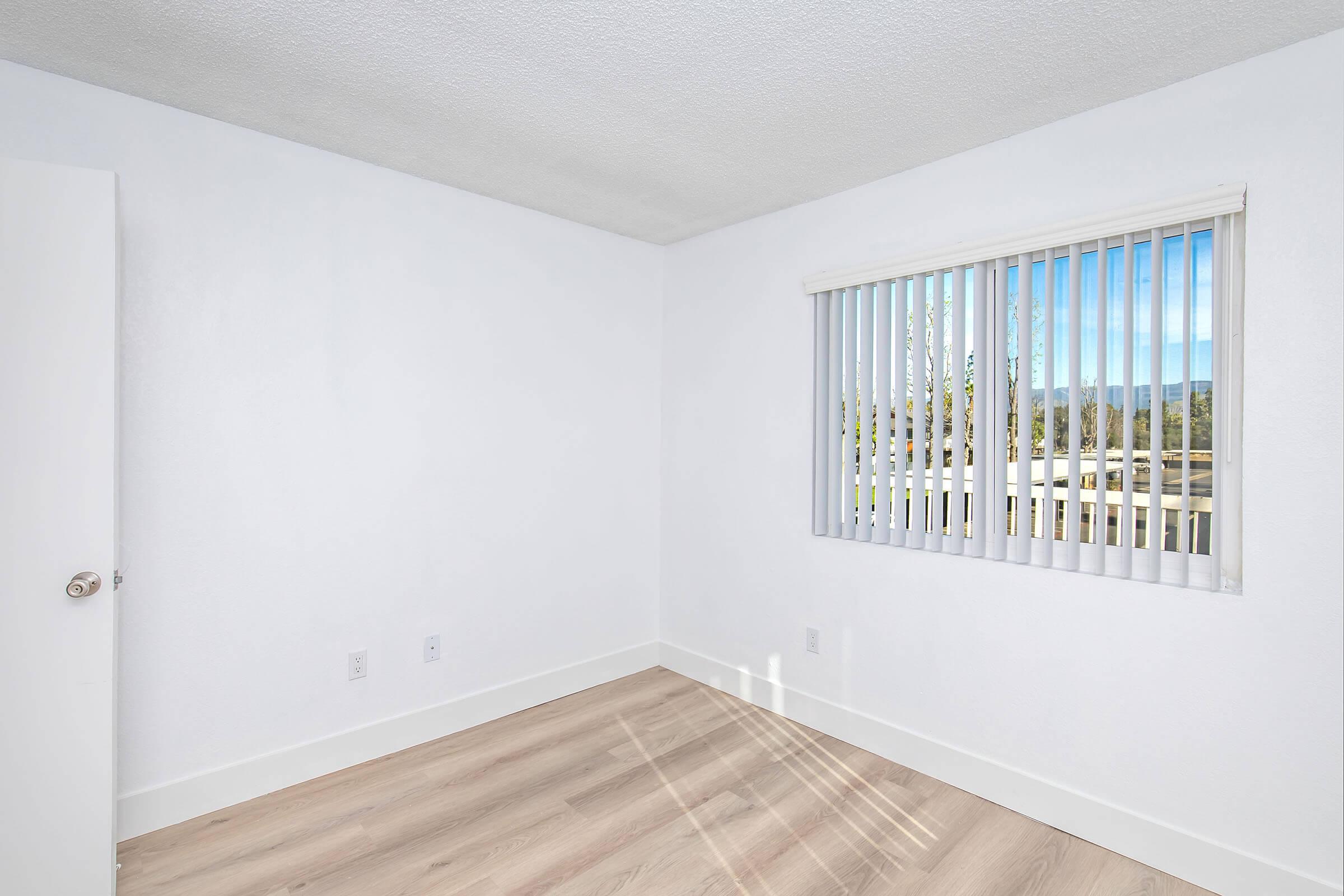 A bright, empty room with white walls and light wooden flooring. A window with vertical blinds allows natural light to enter, creating soft shadows on the floor. The room appears spacious and is ready for furnishing.
