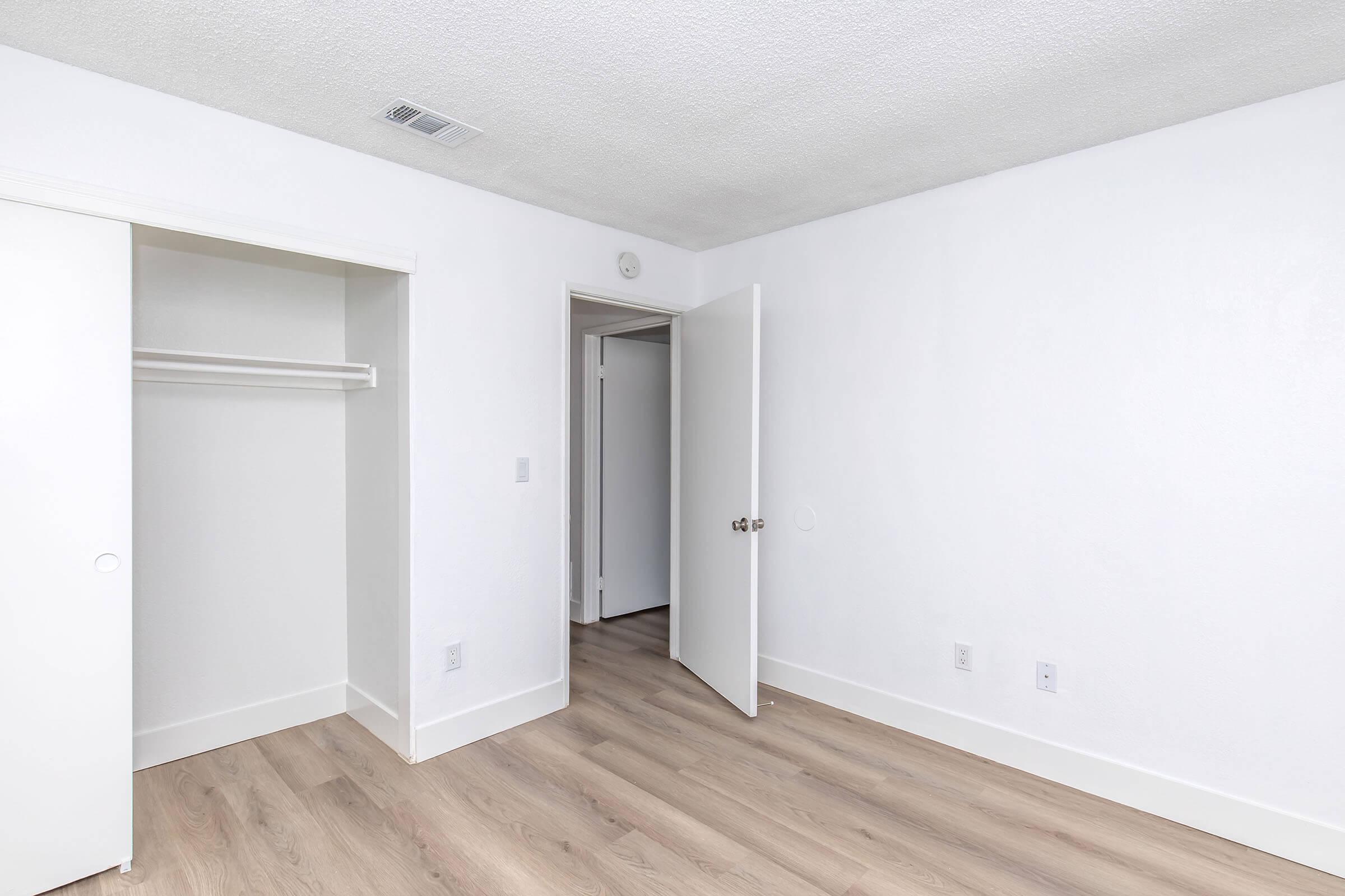 A clean, empty room featuring light wood flooring, white walls, and a small closet with sliding doors. There is a partially open door leading to another room, and the ceiling has a textured finish. Natural light brightens the space, creating a fresh and modern atmosphere.