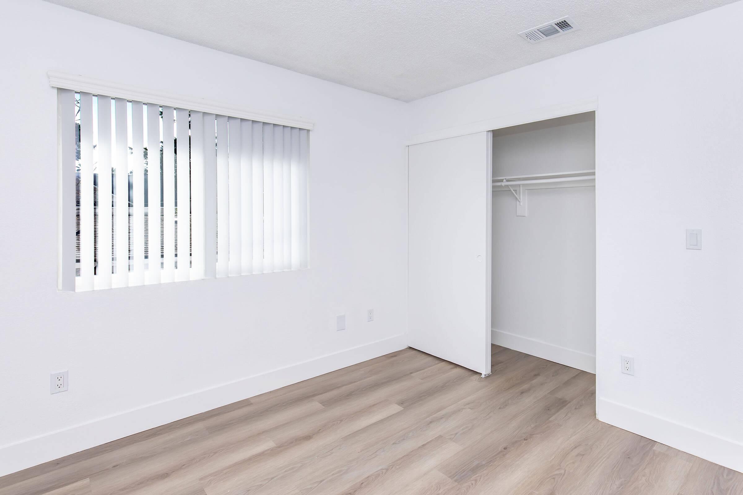 A bright, minimalist room with white walls and light wooden flooring. It features a window with vertical blinds allowing natural light in. There is a closet with sliding doors located on one side, and the overall space appears clean and uncluttered, ideal for various uses such as a bedroom or office.