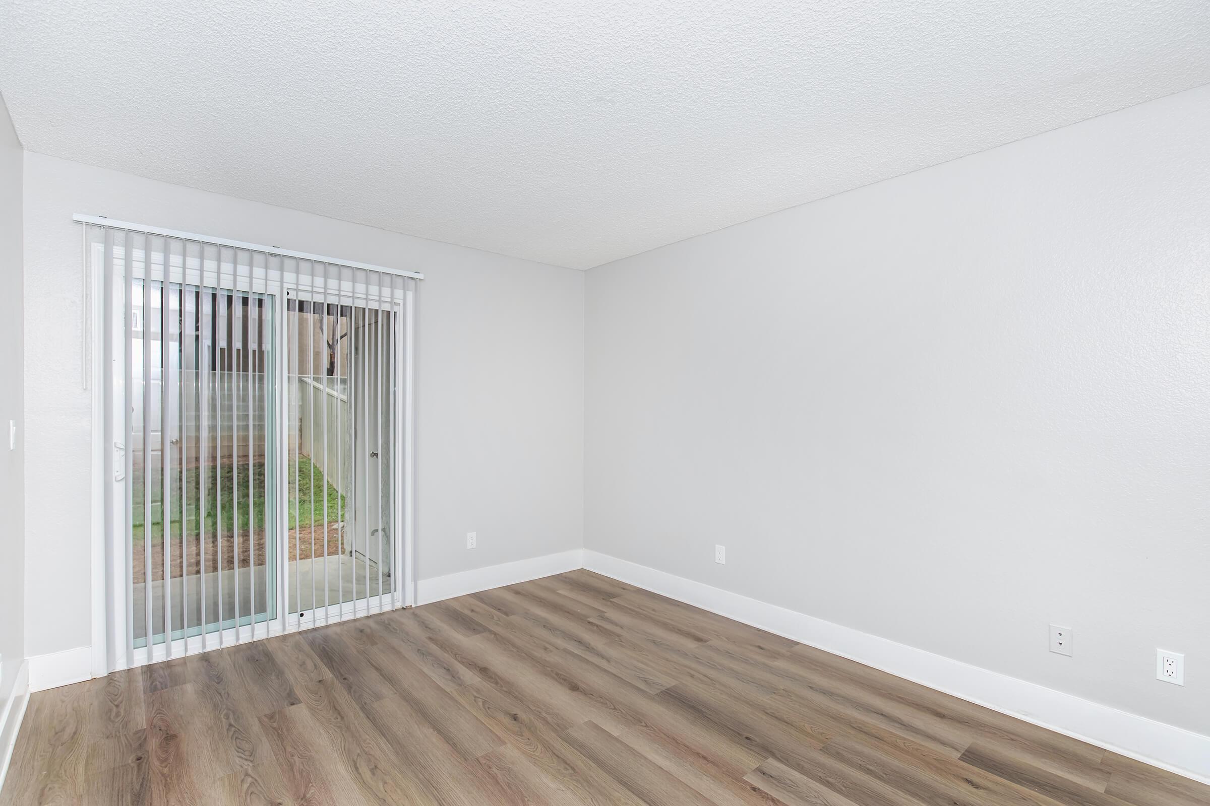 A vacant room with gray walls and a sliding glass door leading to an outdoor area. The floor is finished with light wood laminate. Natural light enters through the door, creating a bright atmosphere. The space is empty and ready for furnishings.
