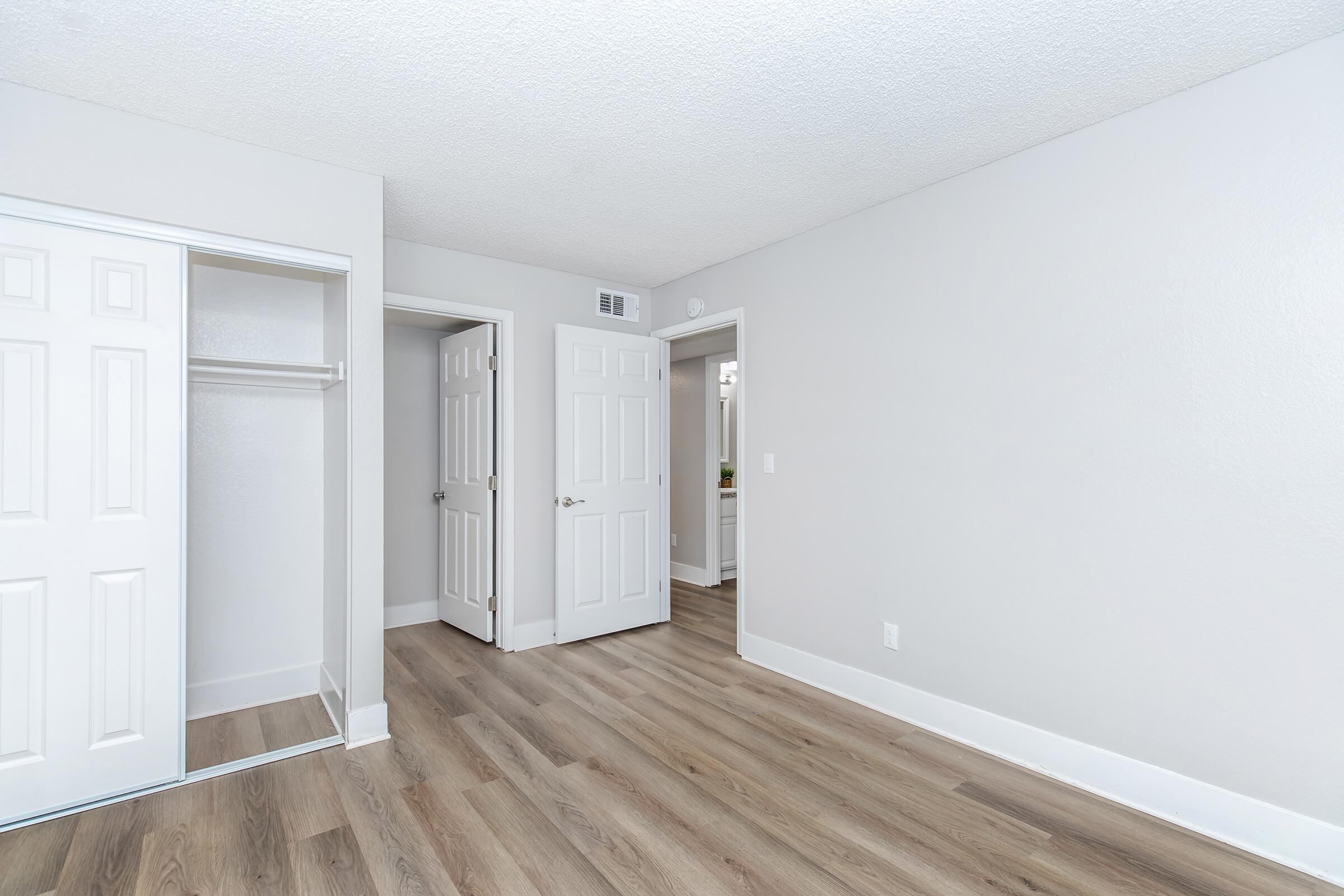 A spacious, light-colored room featuring new hardwood floors. There's a sliding mirror closet on the left, and two white doors leading to other areas. The walls are painted in soft gray, creating a clean and modern aesthetic.