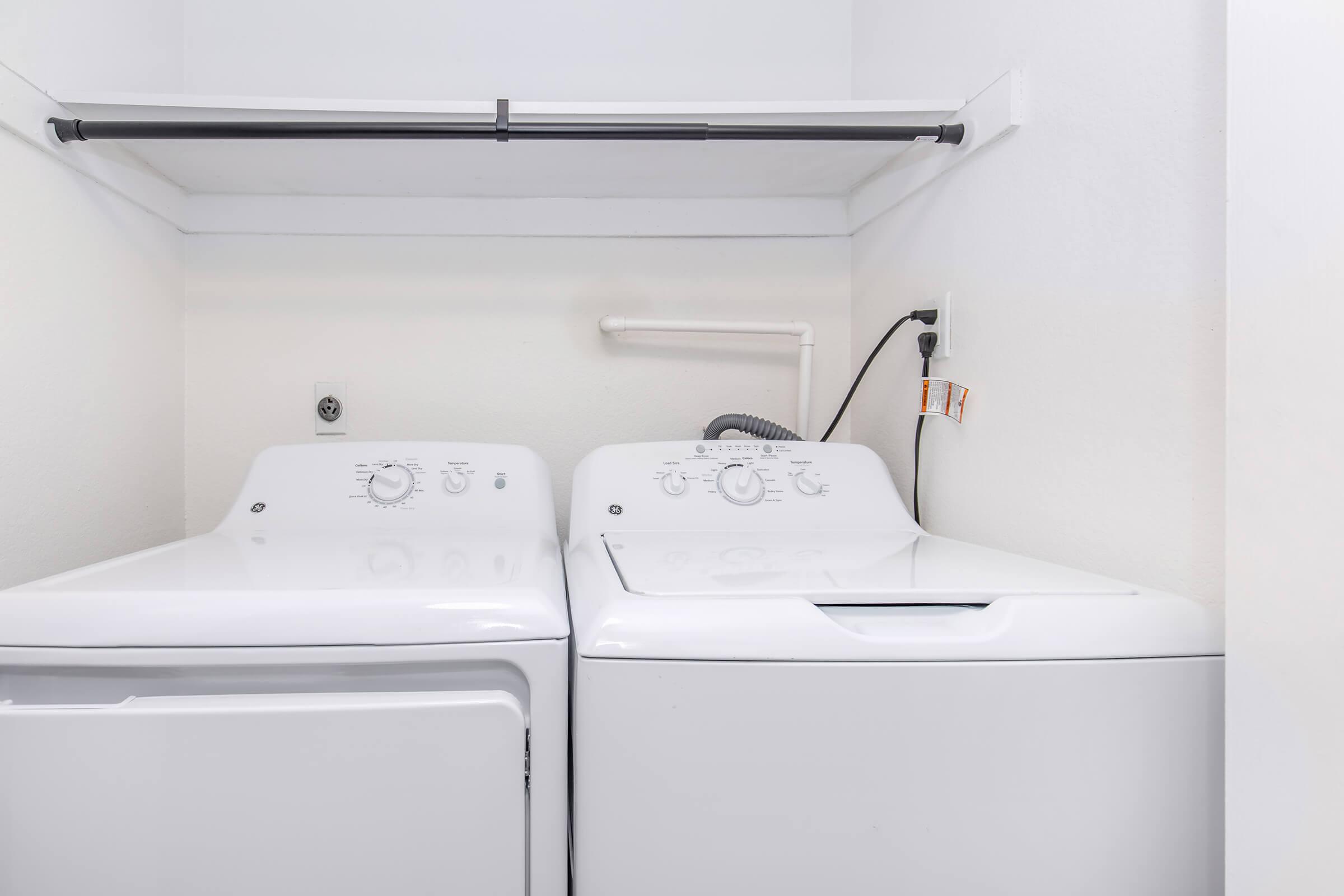 A clean laundry space featuring a stacked washer and dryer against a white wall. The appliances are modern with control knobs and settings clearly visible. A pipe and electrical outlet are also present, contributing to the functional laundry setup.