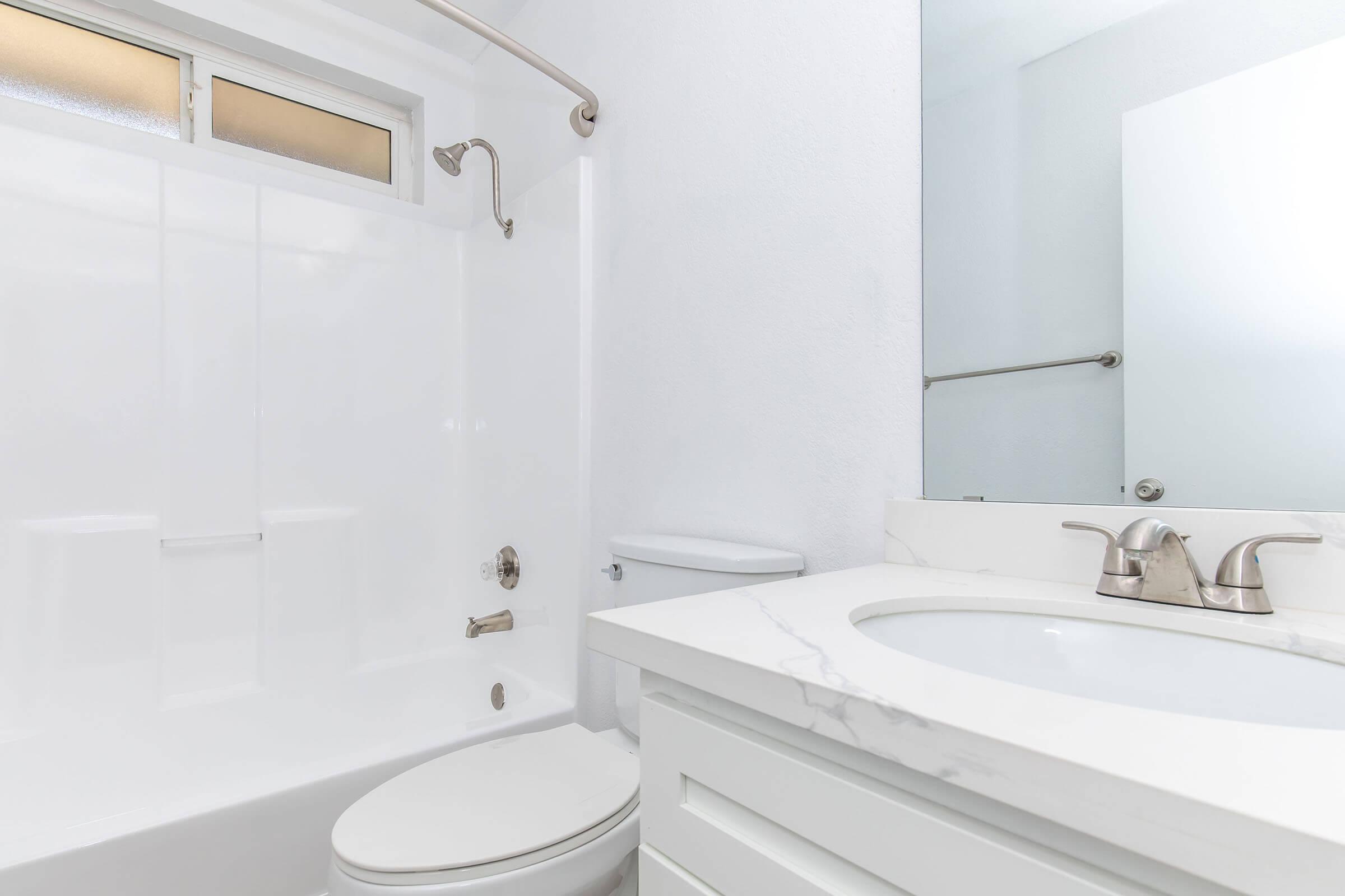 Modern bathroom featuring a white shower tub combo, a sleek toilet, and a marble countertop sink. The walls are painted white, and there is a mirror above the sink. A shower curtain rod is installed, but no curtain is visible. A window allows natural light into the space.