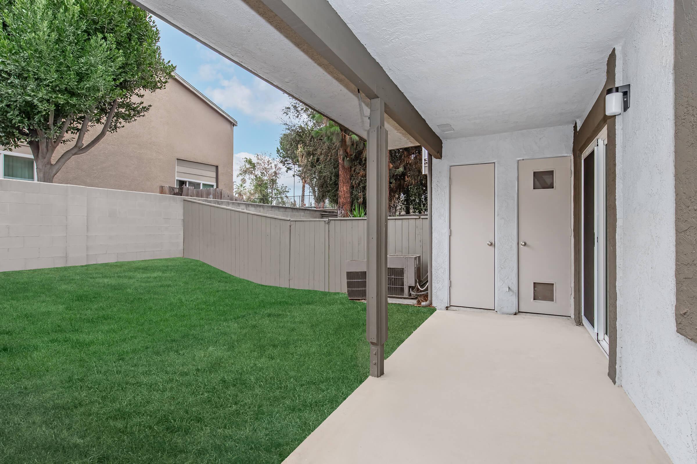 A small covered patio with a light-colored floor overlooks a grassy backyard. The space has a simple design with a door and an adjacent wall, surrounded by trees and nearby houses. The sky is partially cloudy, adding a sense of openness to the area.
