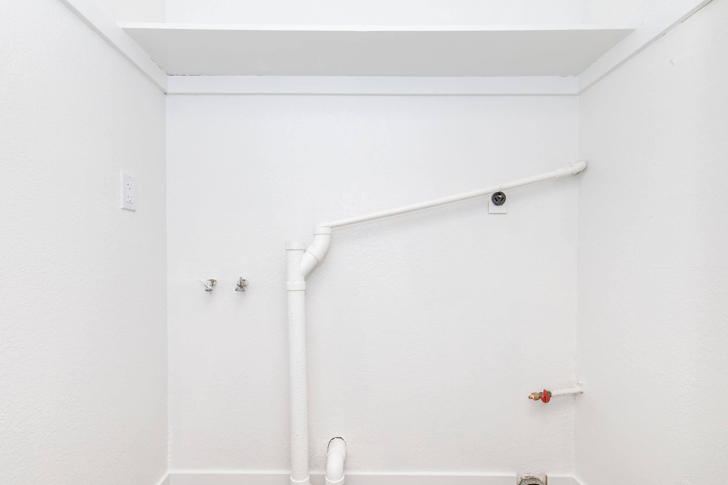 A clean, minimalistic laundry room with white walls. A vertical shelf is mounted above a white pipe system, which connects to two water connections on the wall. The floor is light-colored and the overall space appears bright and uncluttered.