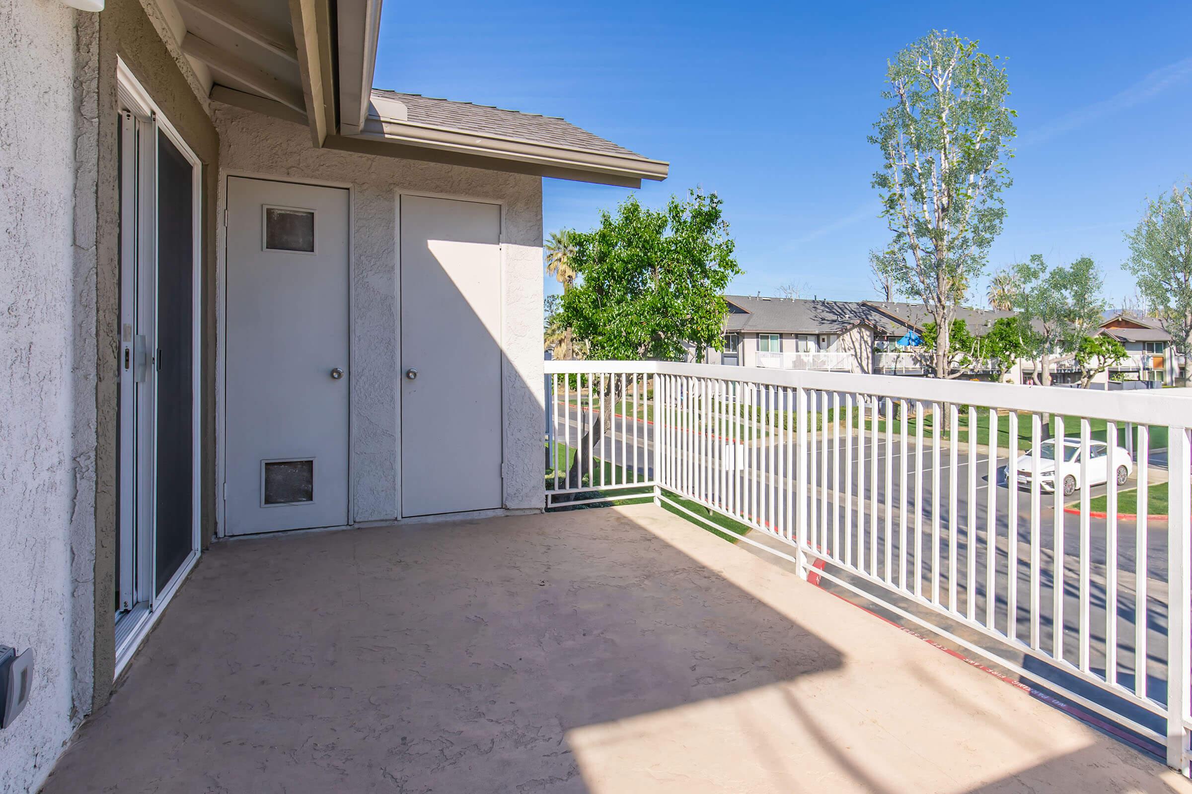 A spacious balcony with a gray concrete floor, surrounded by a white railing. Two closed doors are visible, one with a small dog door. In the background, you can see green trees and parked vehicles in a residential area under a clear blue sky.